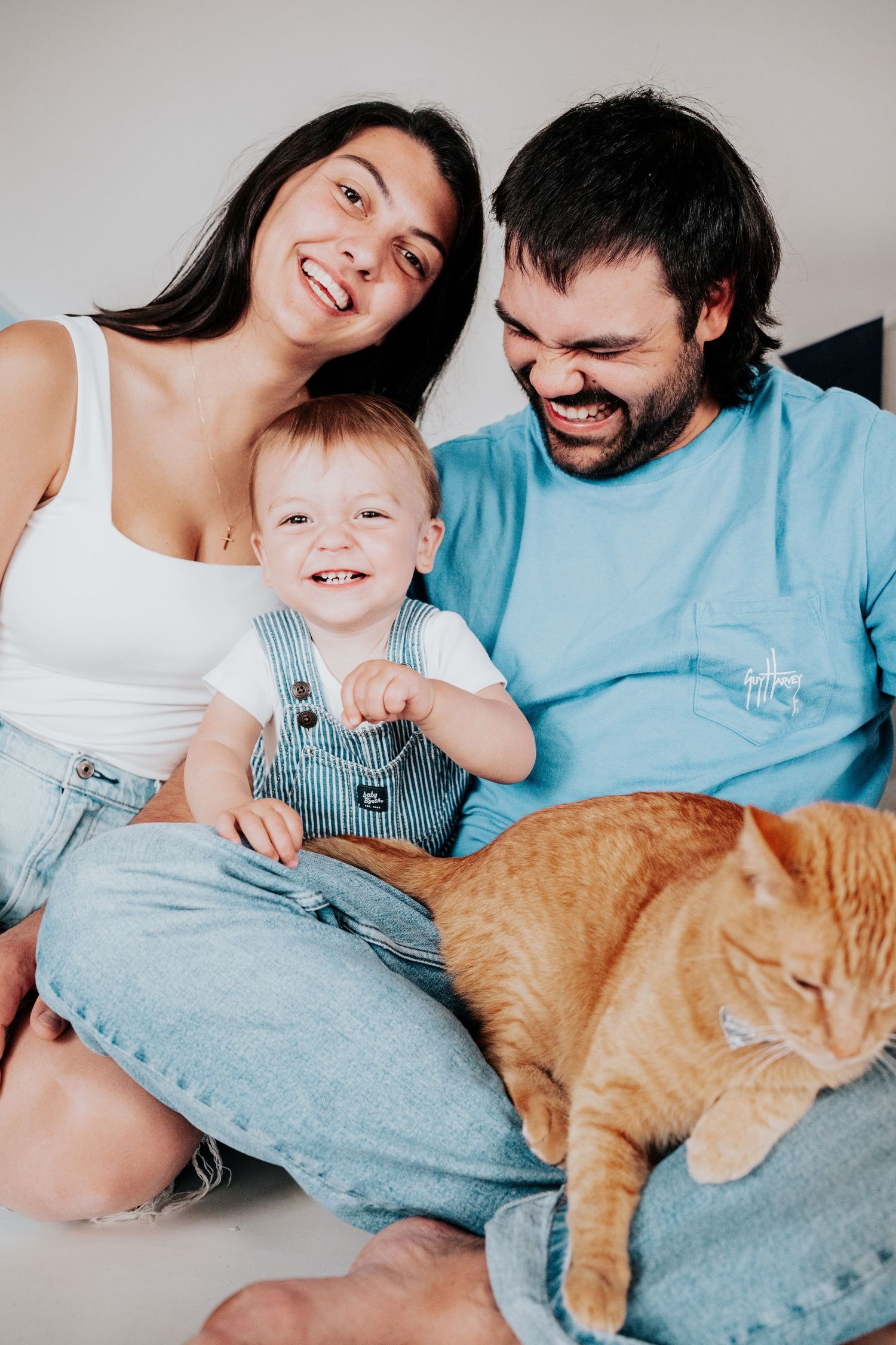 Smiling parents and a cheerful baby in blue overalls sit with a calm orange tabby cat on a white floor.