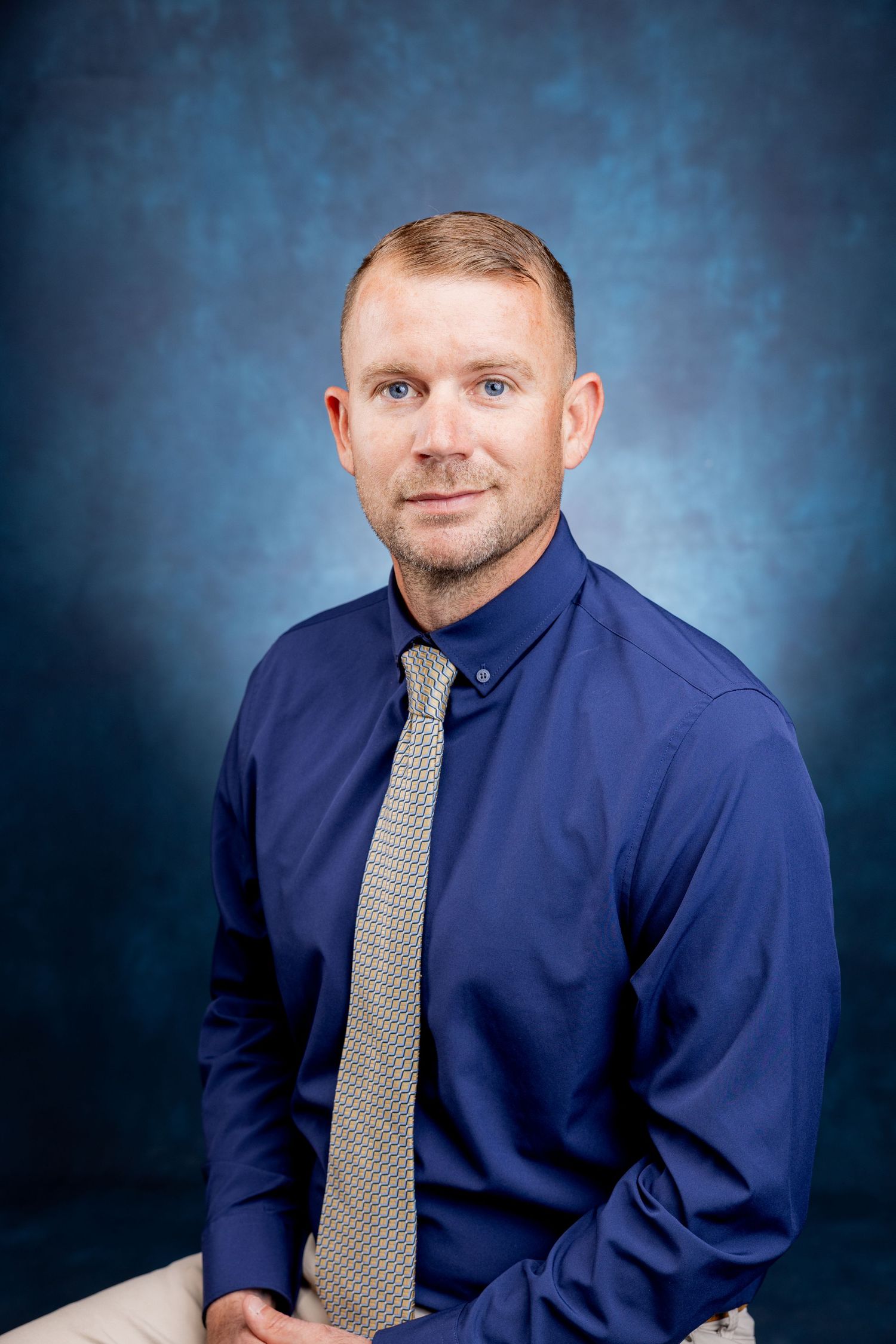 Professional portrait of a man in a navy shirt and gray tie seated casually against a blue background.