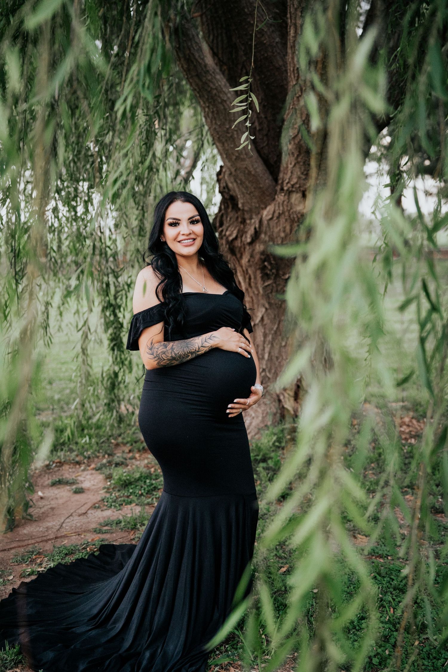 Beautiful pregnant woman in black off-shoulder gown smiles while standing beneath weeping willow branches.