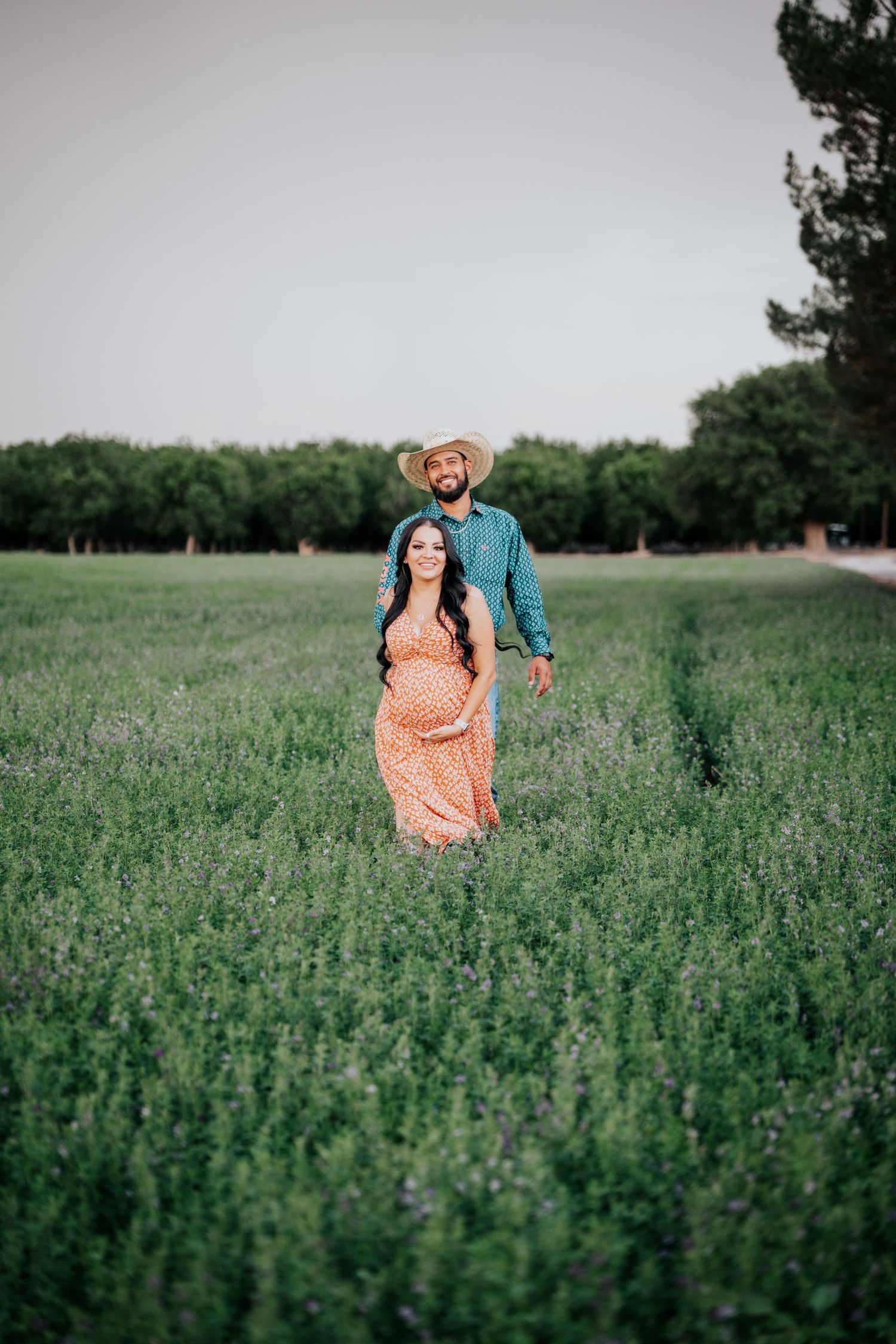 Pregnant couple standing in a lush green field, woman in orange floral dress, man in teal shirt and cowboy hat.