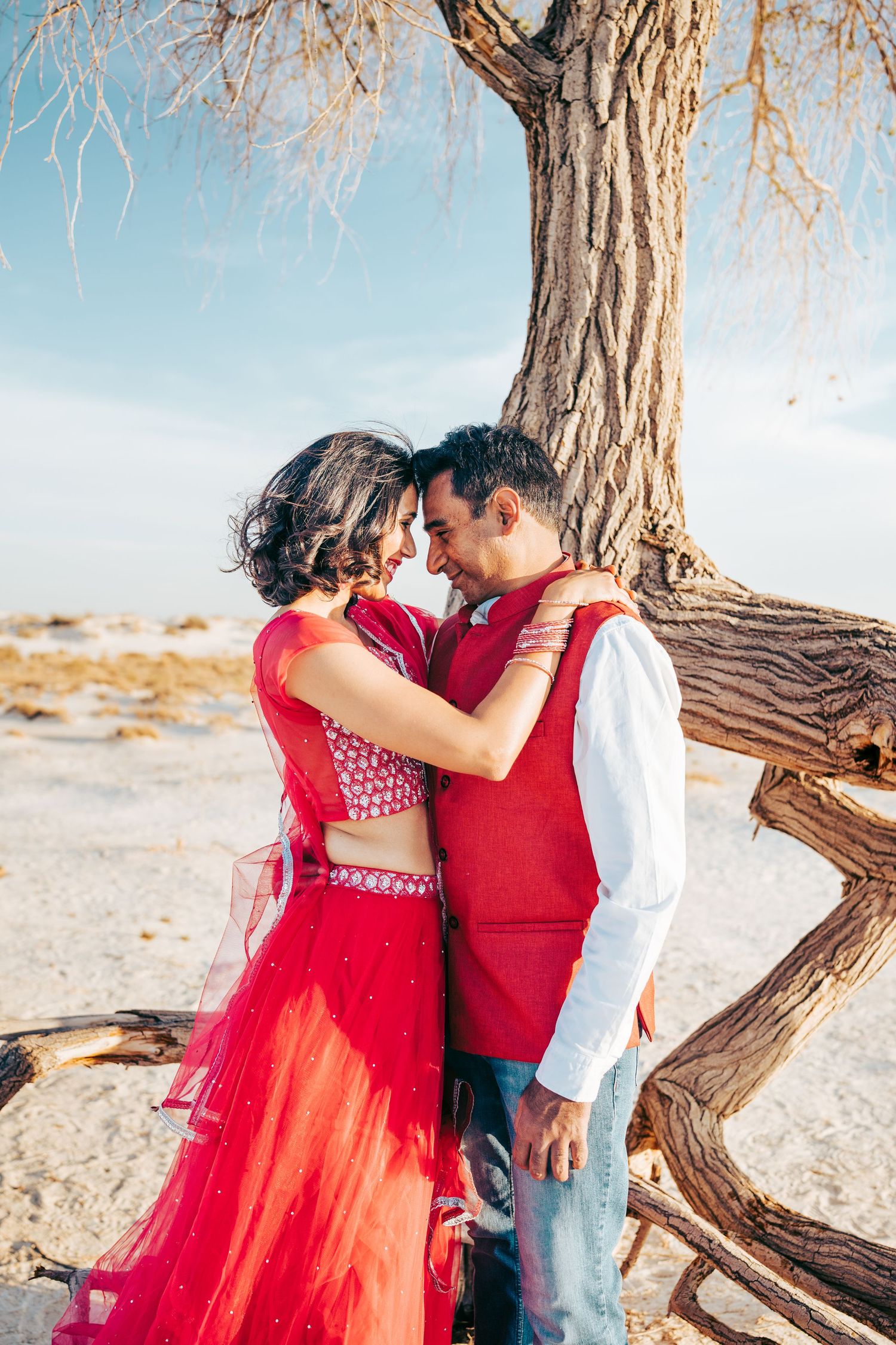 Indian couple in matching red outfits sharing an intimate forehead touch under a desert tree.