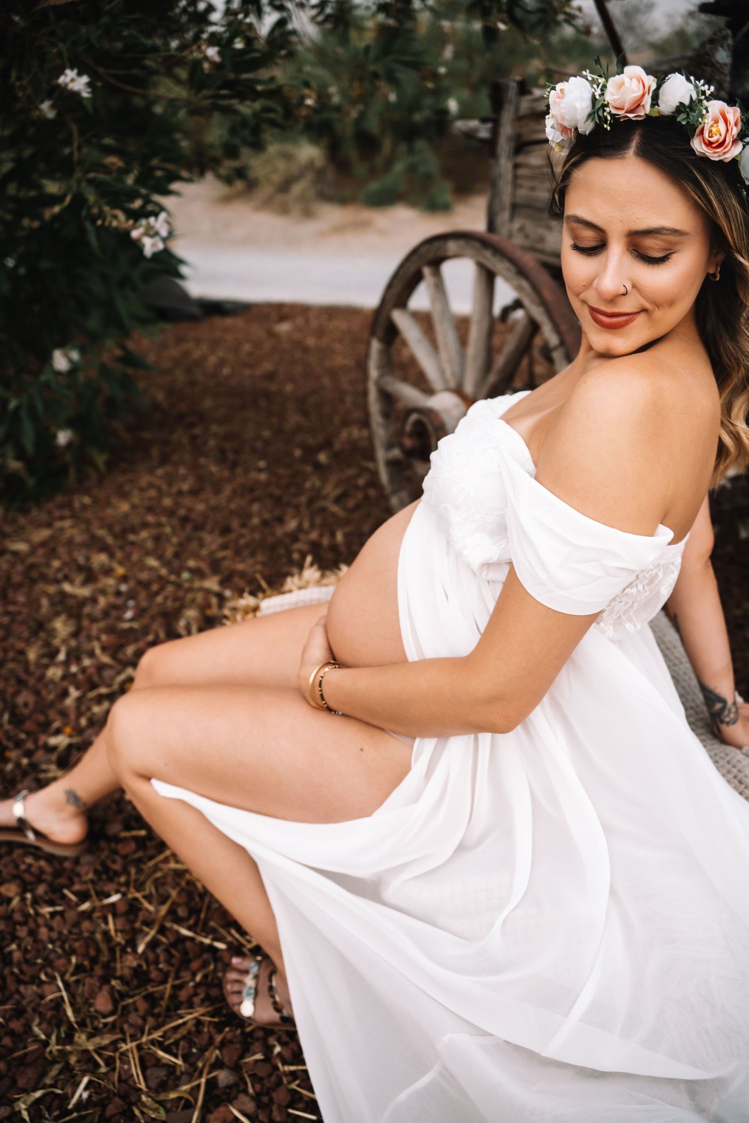 Radiant pregnant woman wearing floral crown and white dress sits outdoors near rustic wooden wheel.