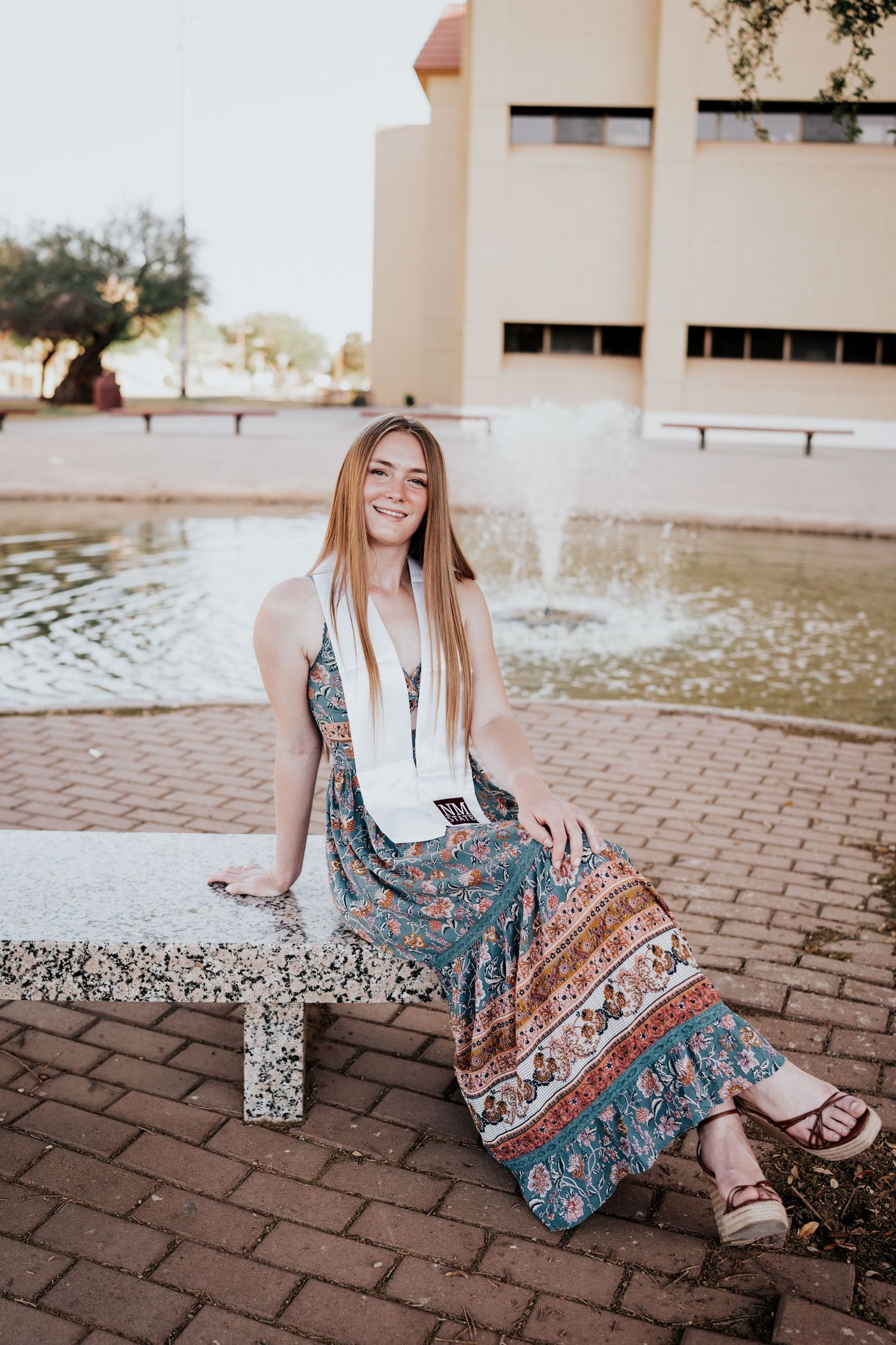 Cheerful young woman in bohemian floral outfit smiling while sitting on bench by fountain.