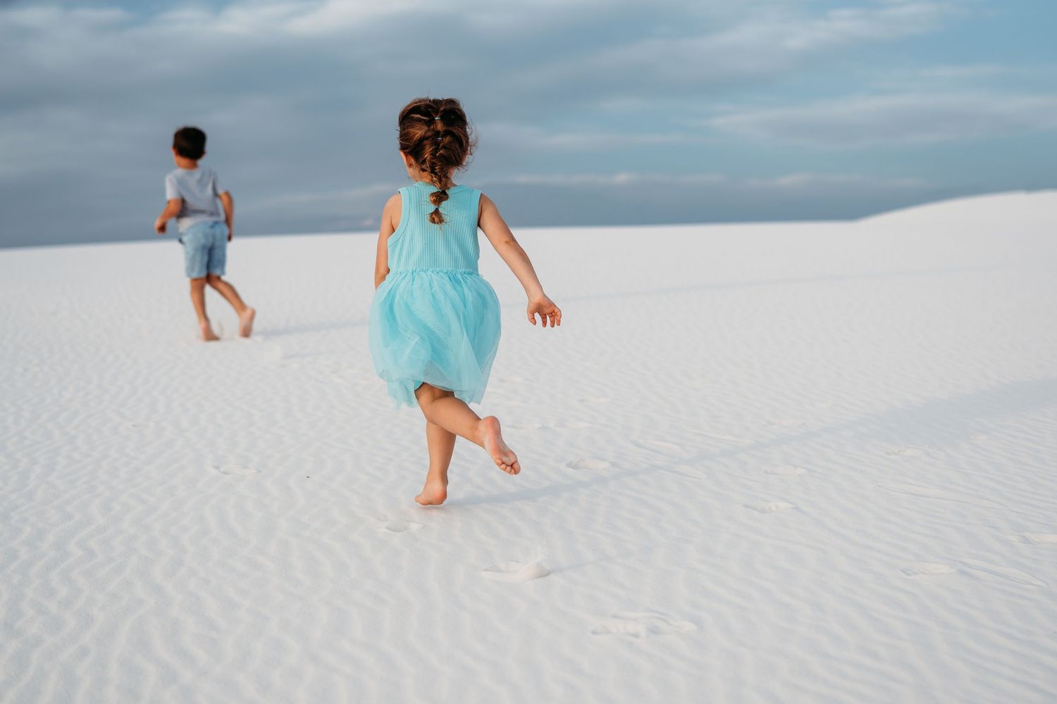 A young girl in a turquoise dress runs barefoot across white sand dunes chasing her brother.