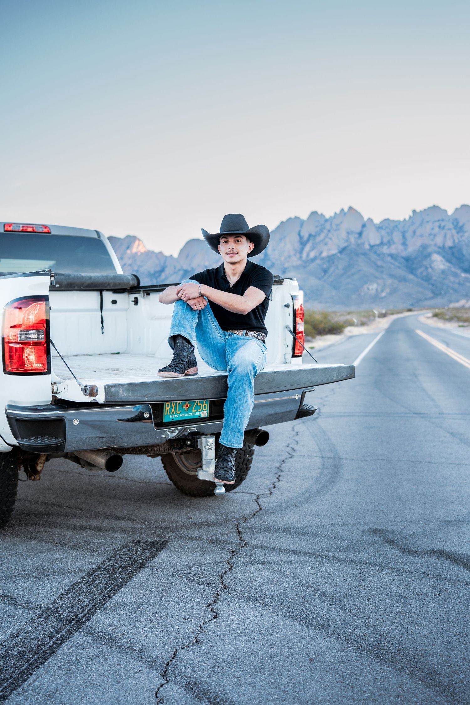 Young cowboy in black hat sits on tailgate of white pickup truck on desert highway with mountains.