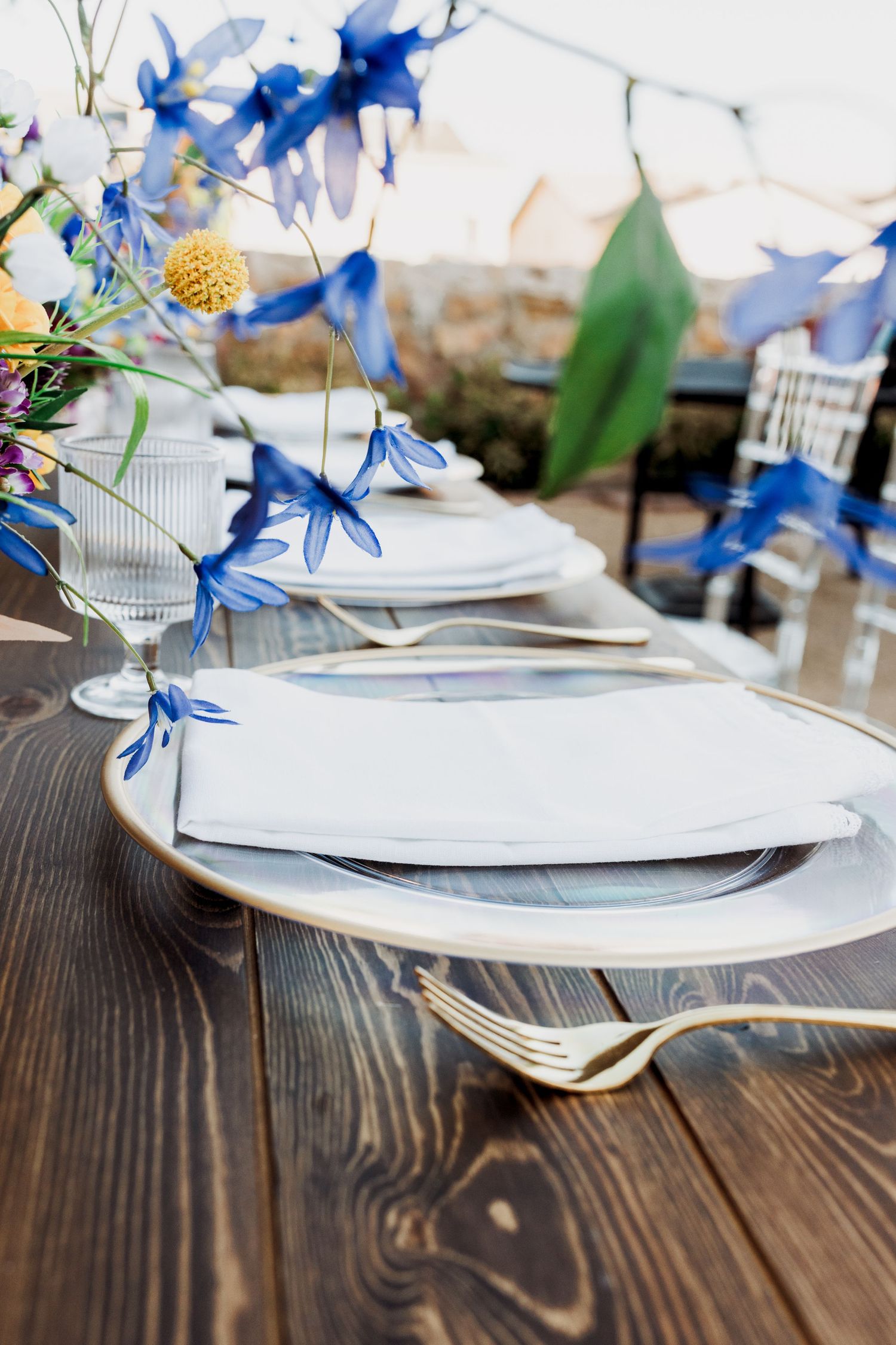 Close-up of place setting on dark wood table with blue delphinium flowers and gold fork in foreground.