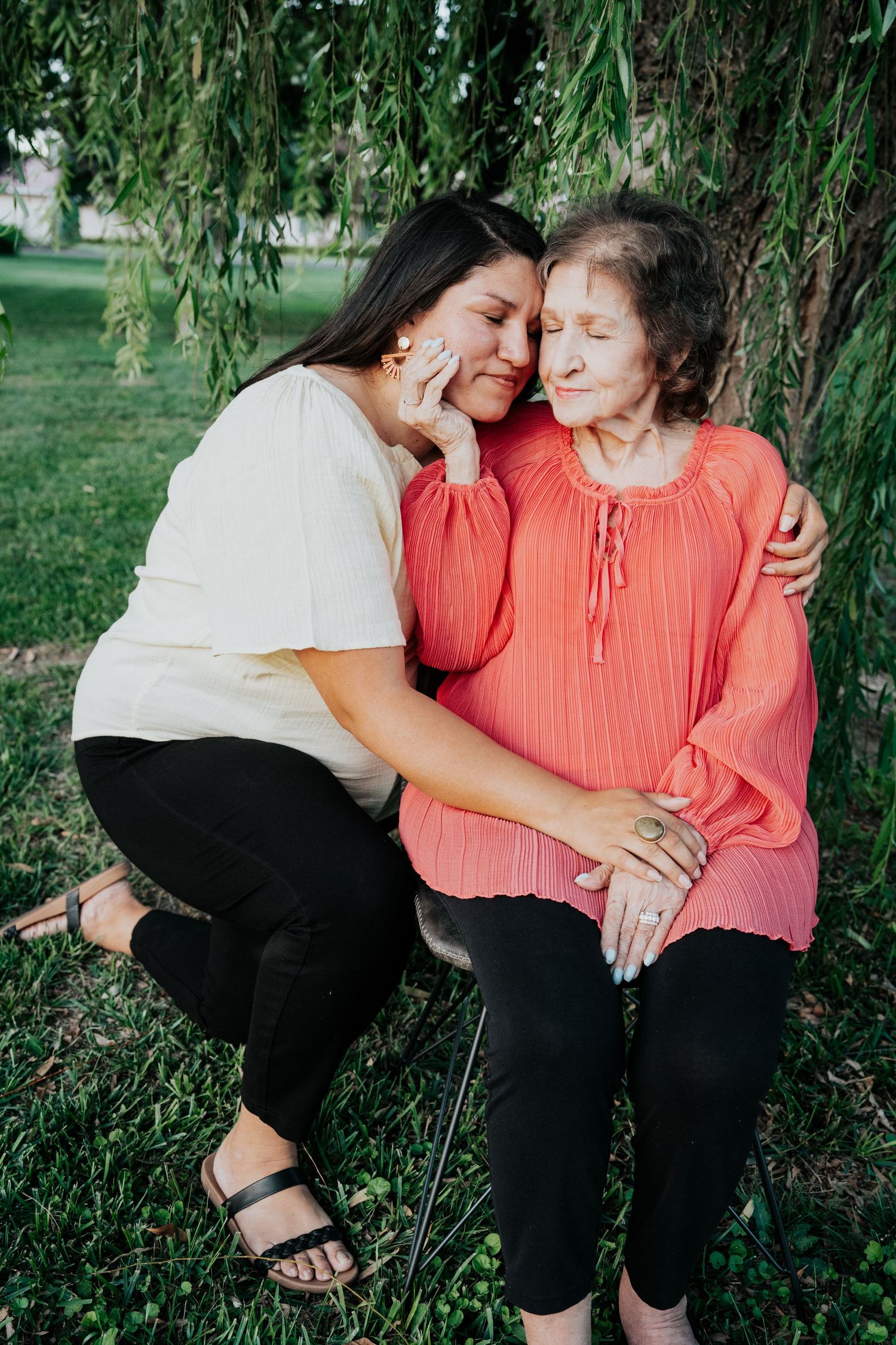 Young woman crying while elderly woman in coral blouse comforts her under a willow tree outdoors.