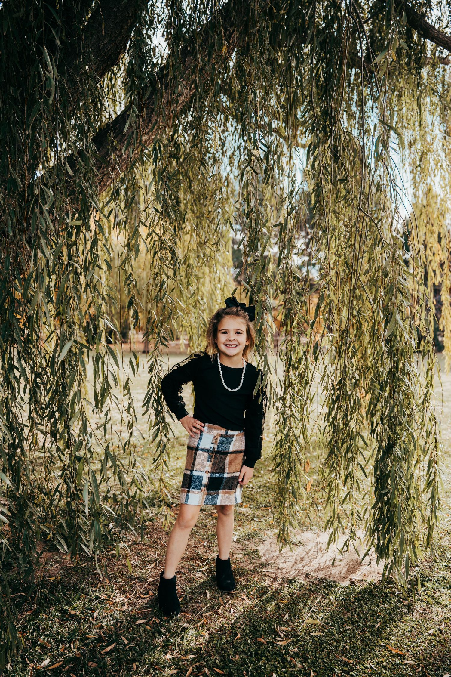 Smiling girl in plaid skirt and black top posing under weeping willow branches by a pond.