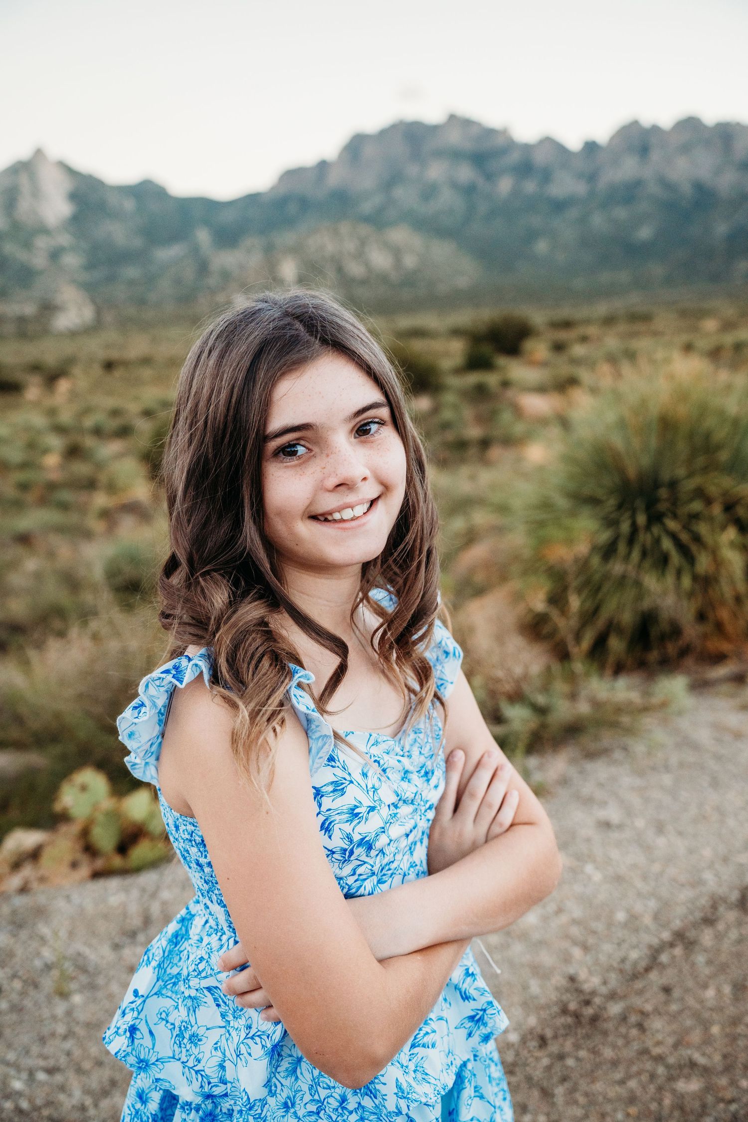 Smiling teenage girl in a blue floral dress standing with arms crossed in a desert landscape.
