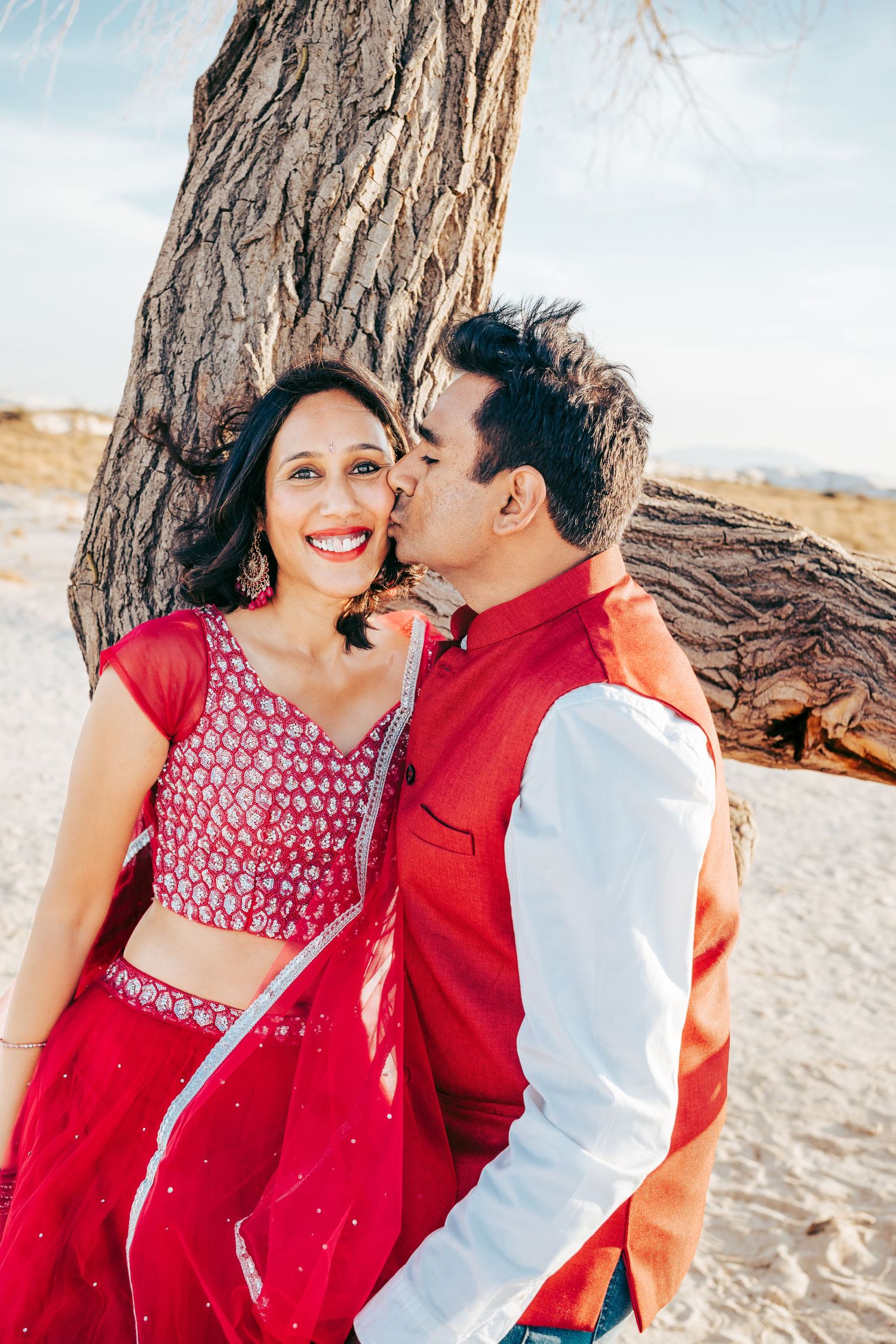 Man in red vest kissing woman in red lehenga on the cheek near a large tree on sandy terrain.