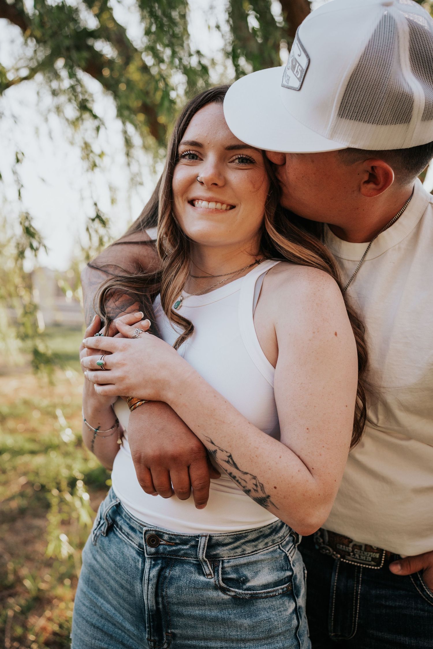 Man in white cap kissing tattooed woman on cheek as she smiles happily in warm outdoor setting.