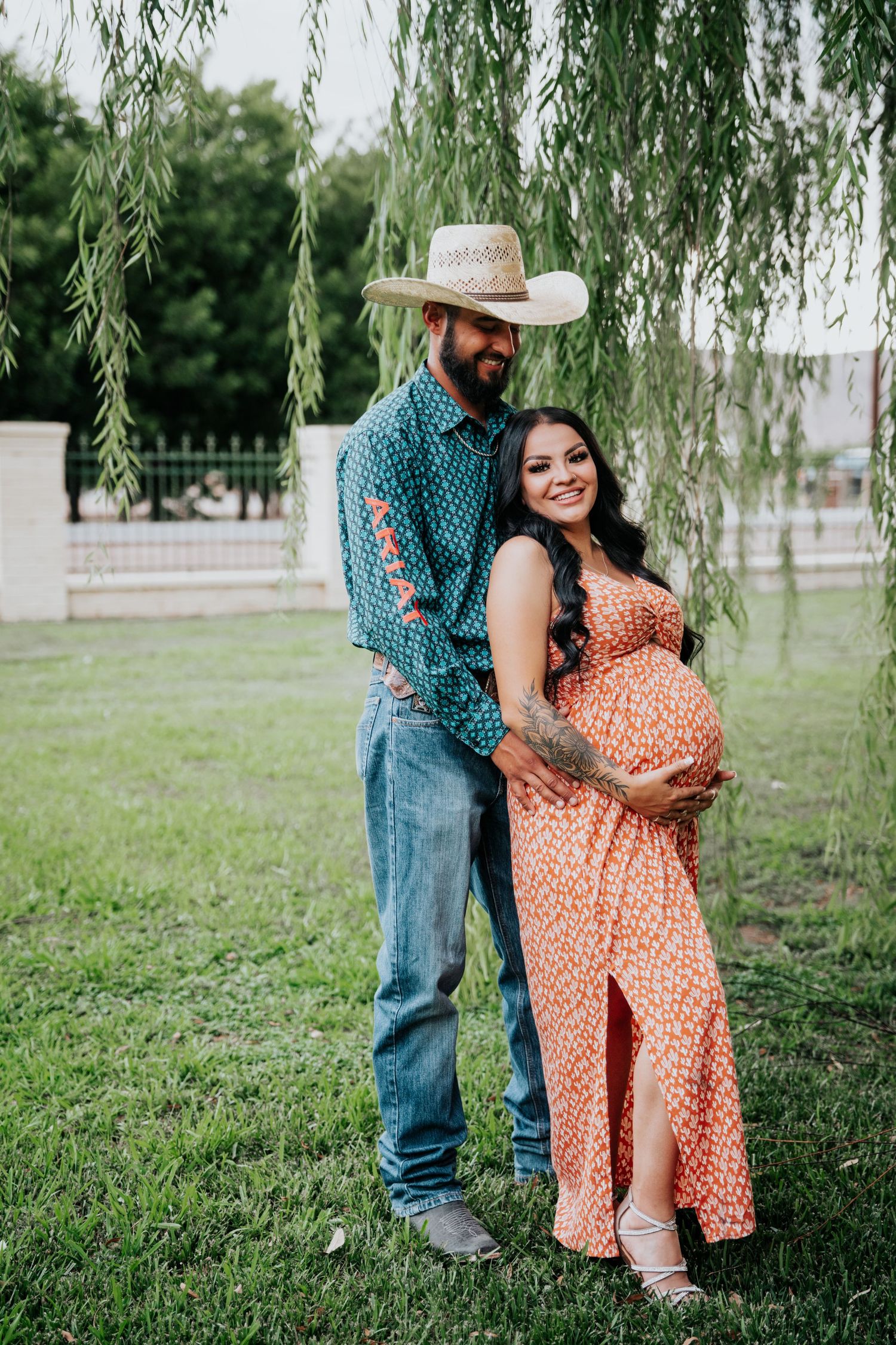 Pregnant couple posing under weeping willow, man in cowboy hat and teal shirt embracing woman in orange floral dress.