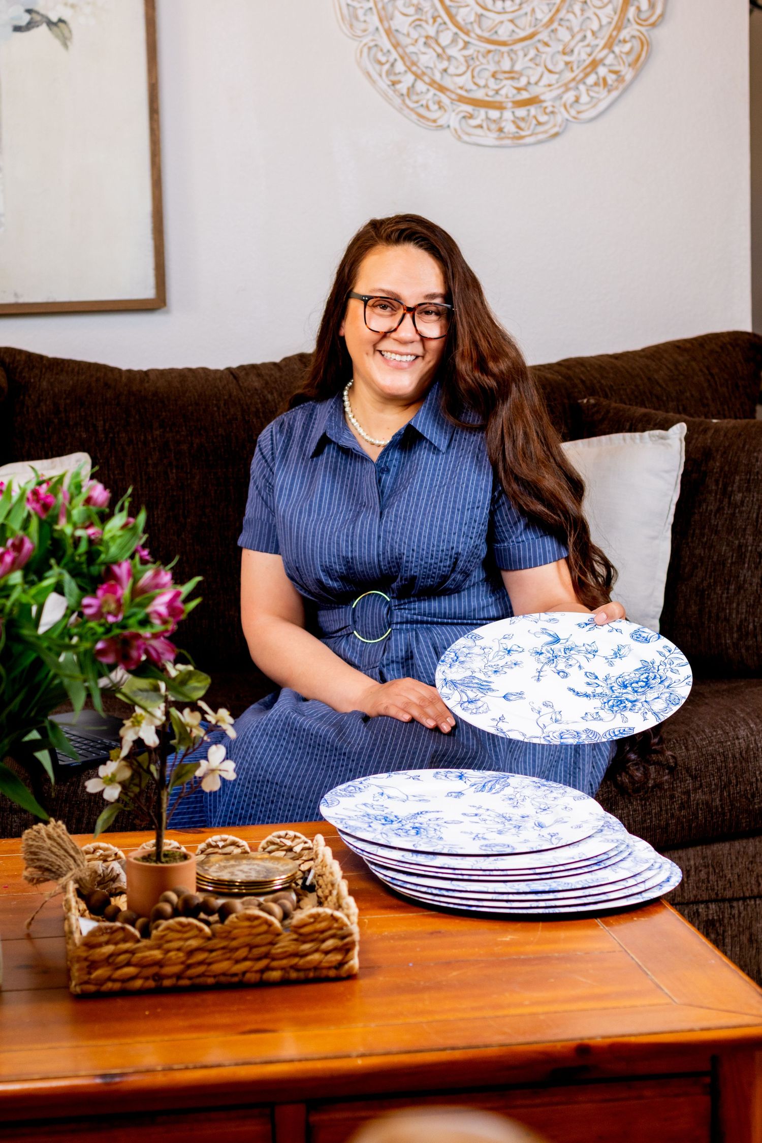 Smiling woman in blue dress sitting on sofa holding a blue and white decorative plate indoors.