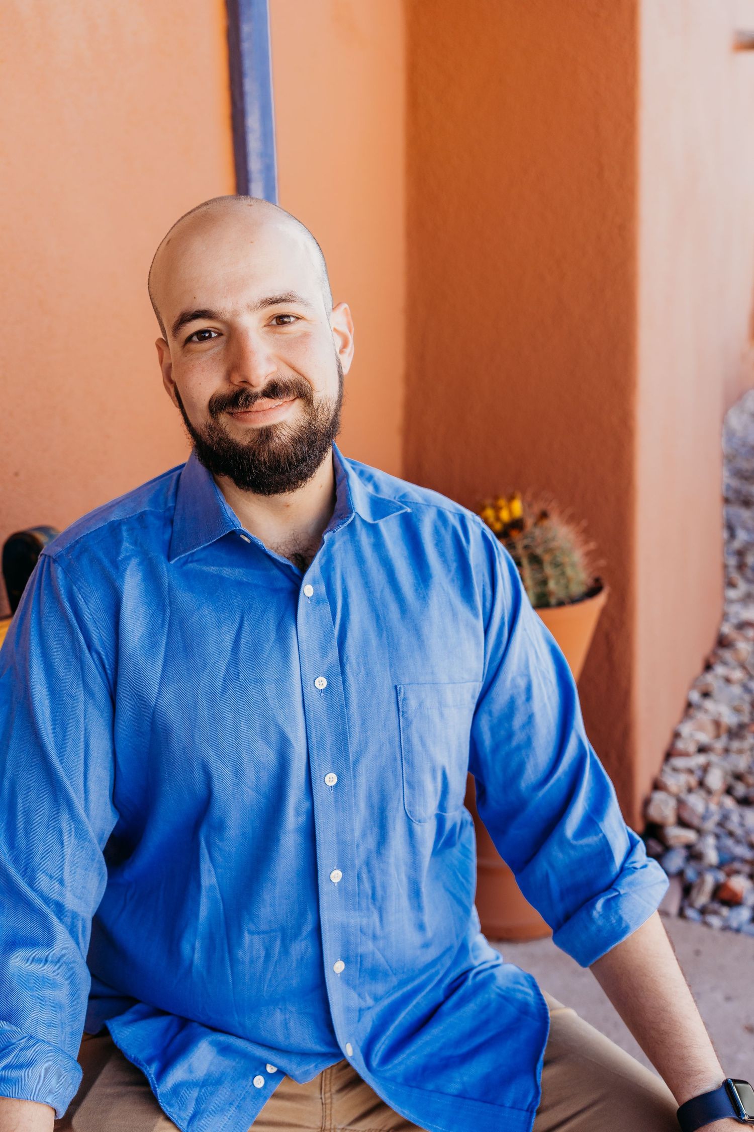 Bald man with beard smiling warmly in blue shirt against orange adobe wall.
