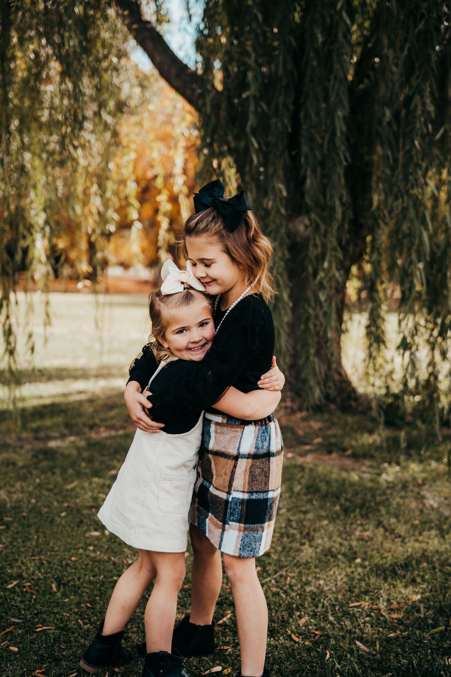 Two young sisters share a warm hug under a weeping willow in autumn park.