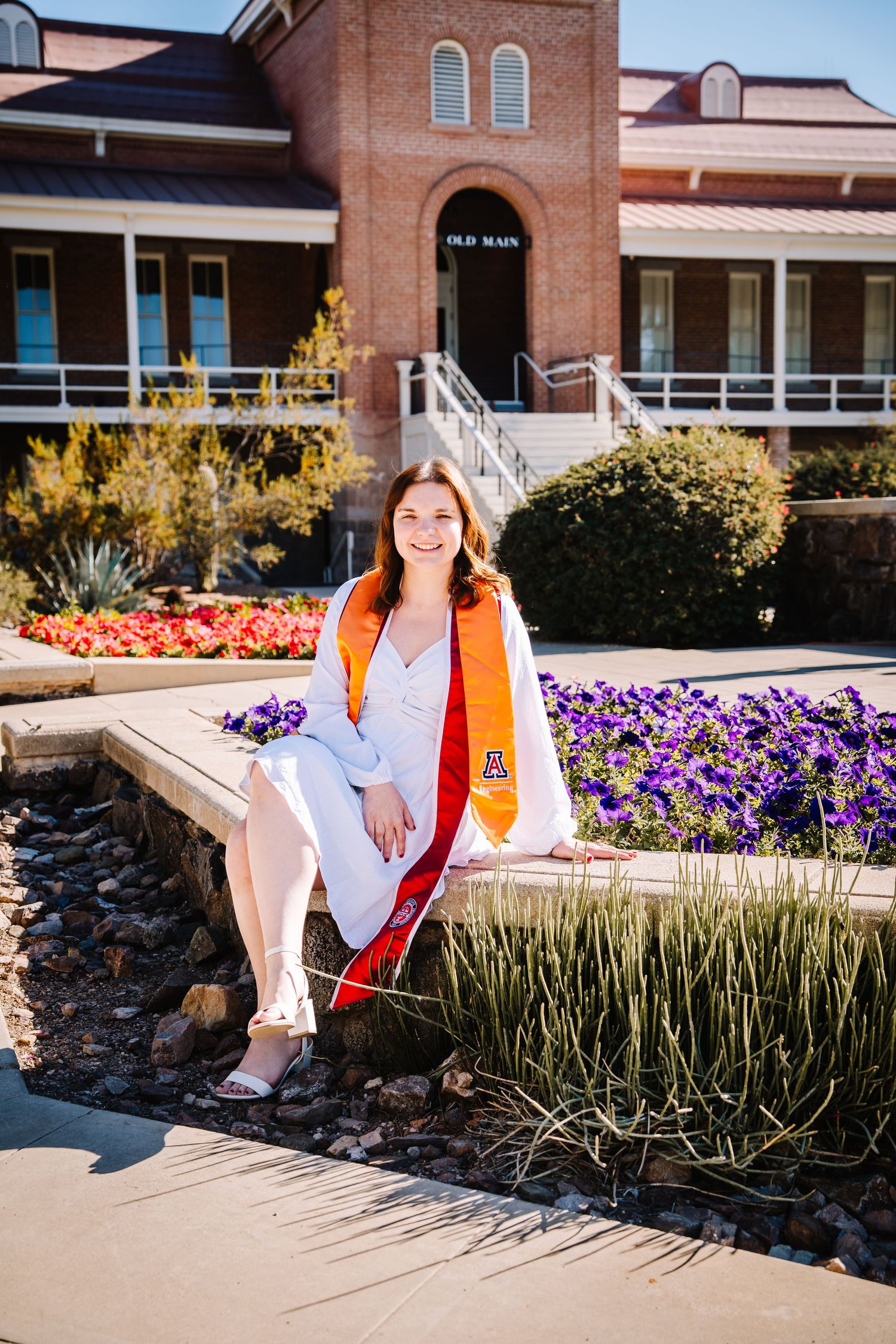 Smiling female graduate in white dress with orange sash sits by colorful garden at Old Main.