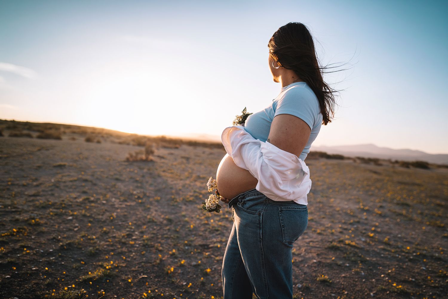 Side profile of pregnant woman cradling belly holding flowers at desert sunset.