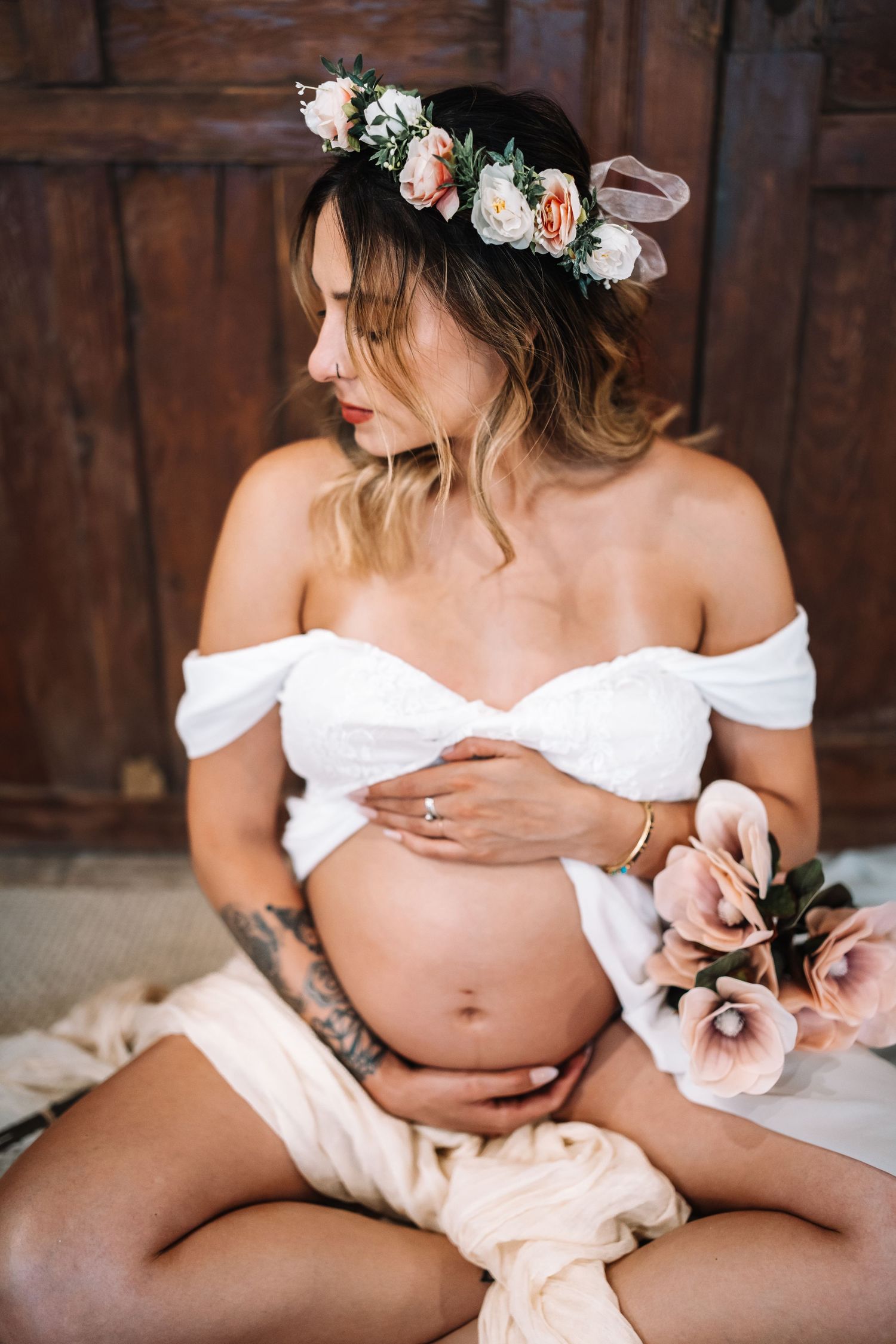 Elegant pregnant woman with flower crown and tattoo holds baby bump beside blush pink flowers in maternity shoot.