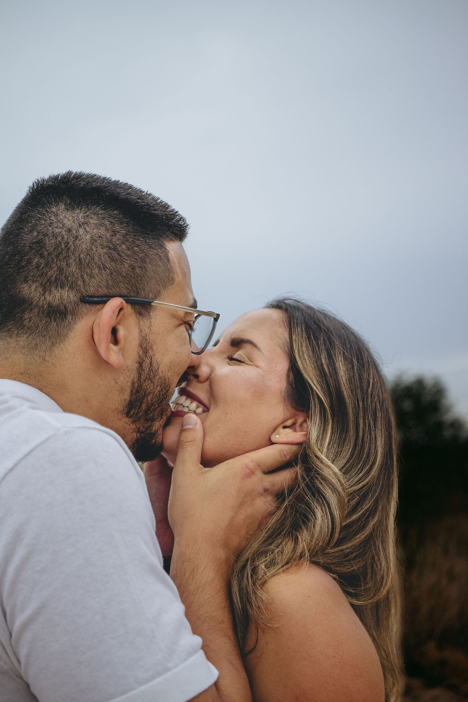Couple sharing a playful kiss outdoors with overcast sky in the background.