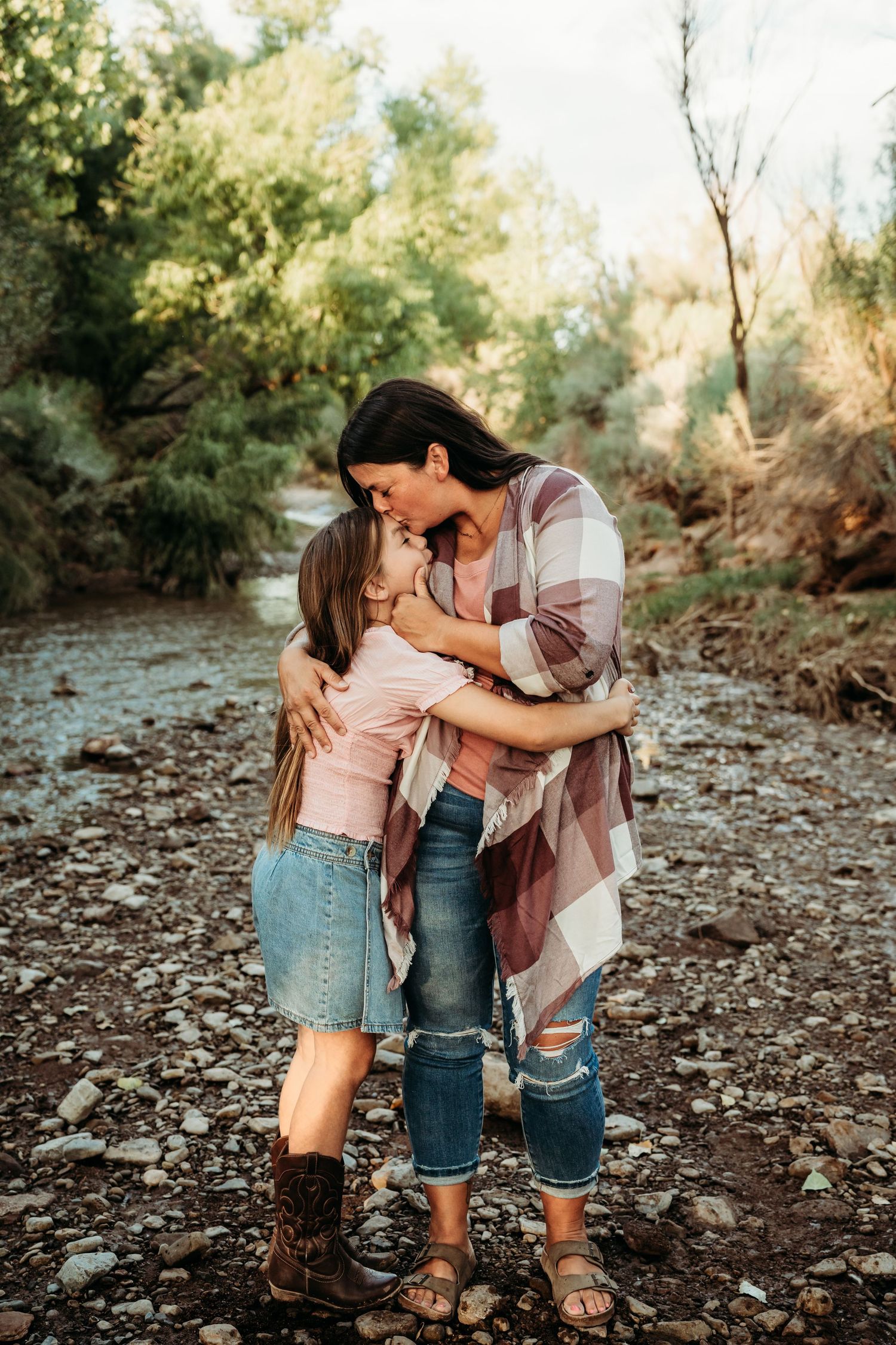 A mother kisses her daughter on the forehead while embracing near a rocky creek in a wooded area.