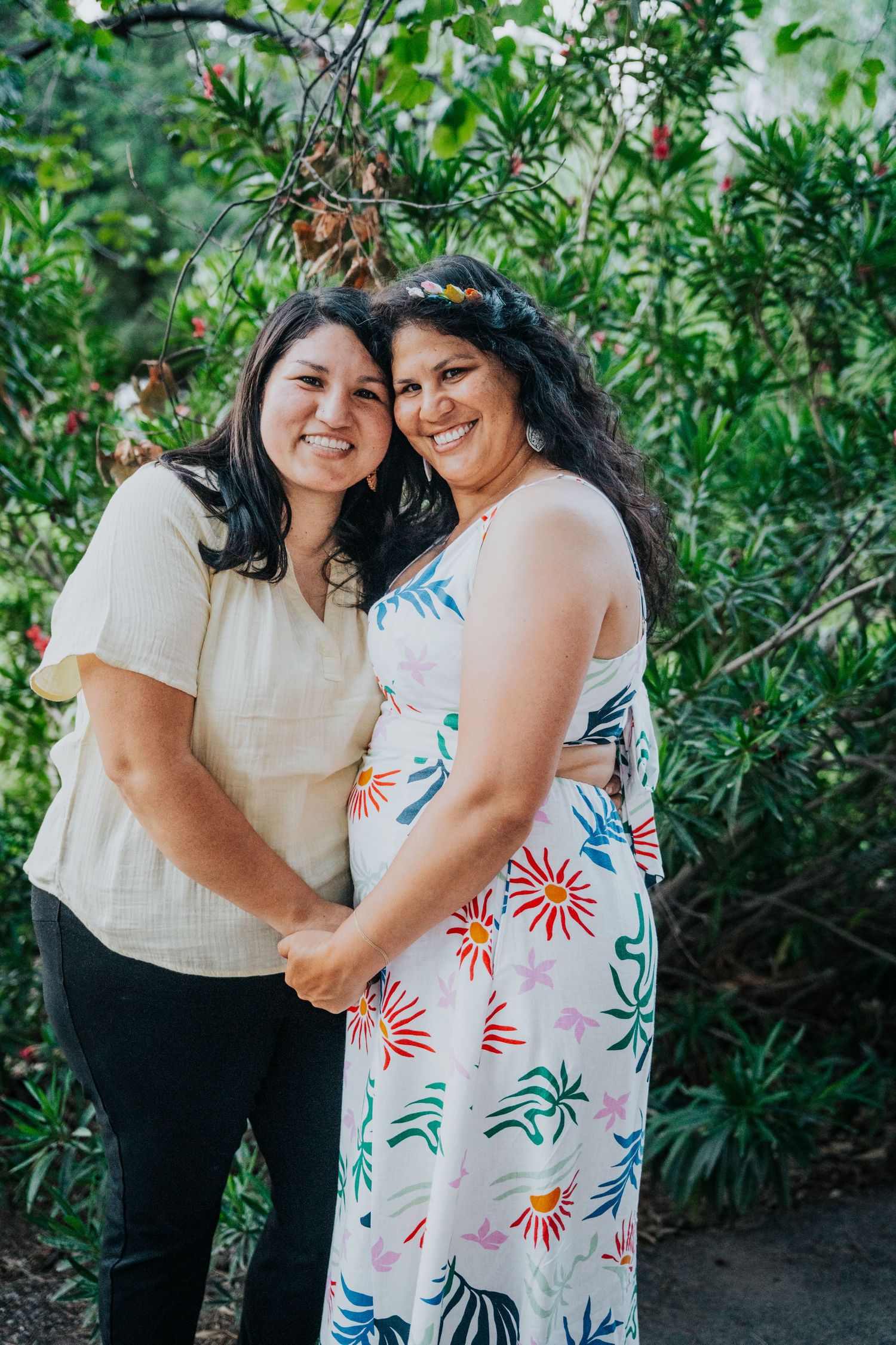 Two smiling women with dark hair embrace warmly in front of lush green foliage outdoors.