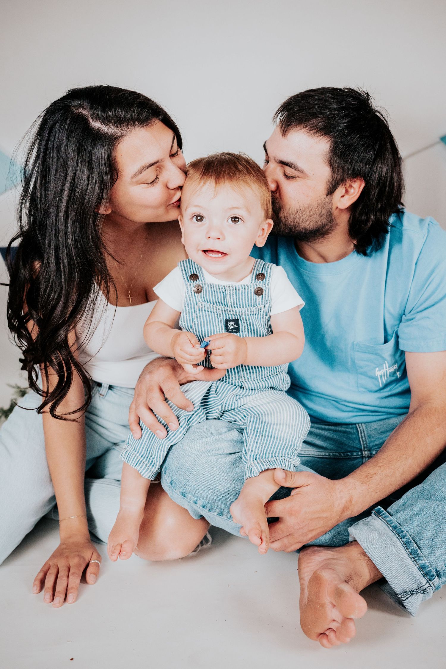 Happy parents kissing their baby boy on cheeks while sitting together on white floor.