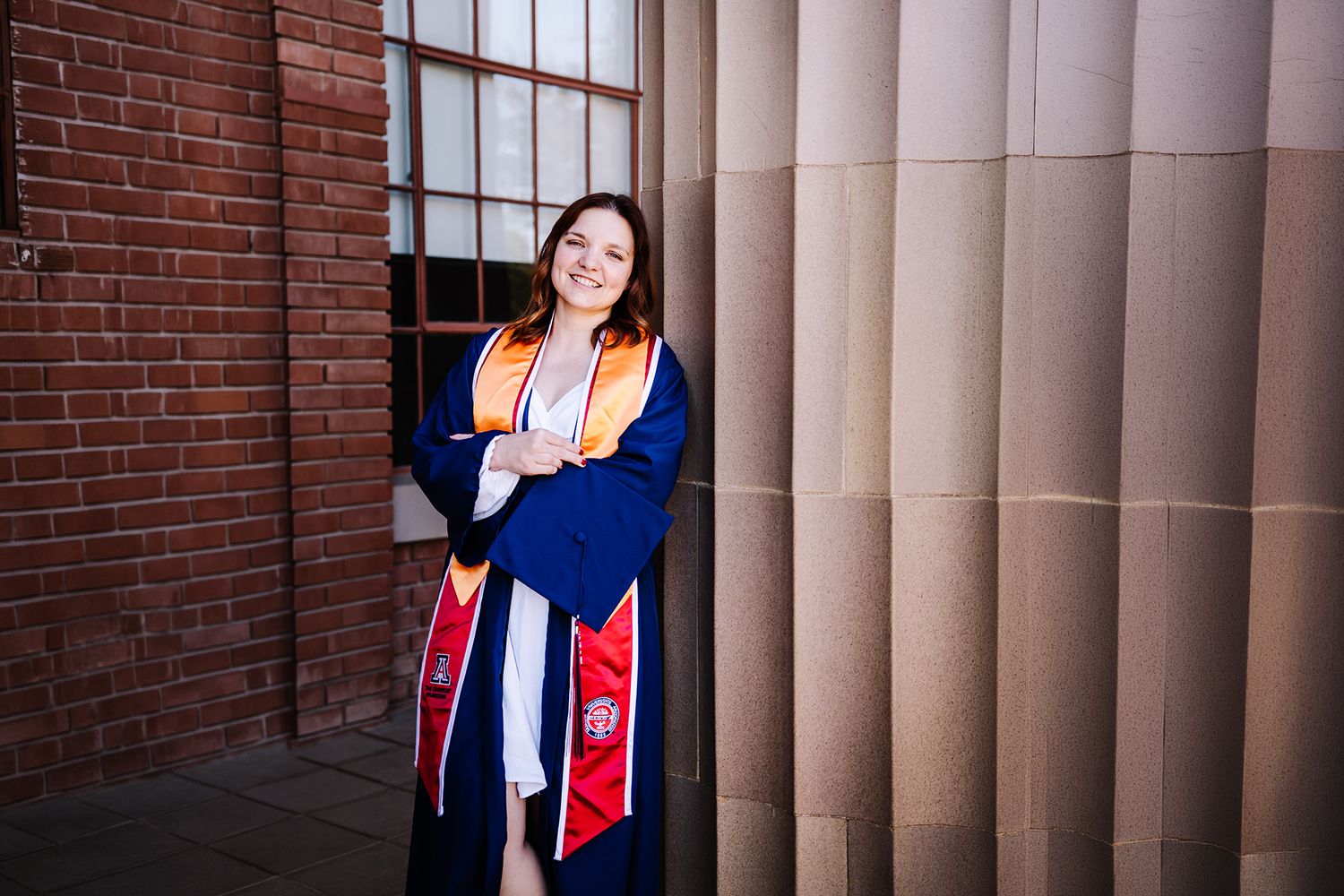 A smiling graduate in blue and orange regalia poses between brick wall and stone columns outdoors.