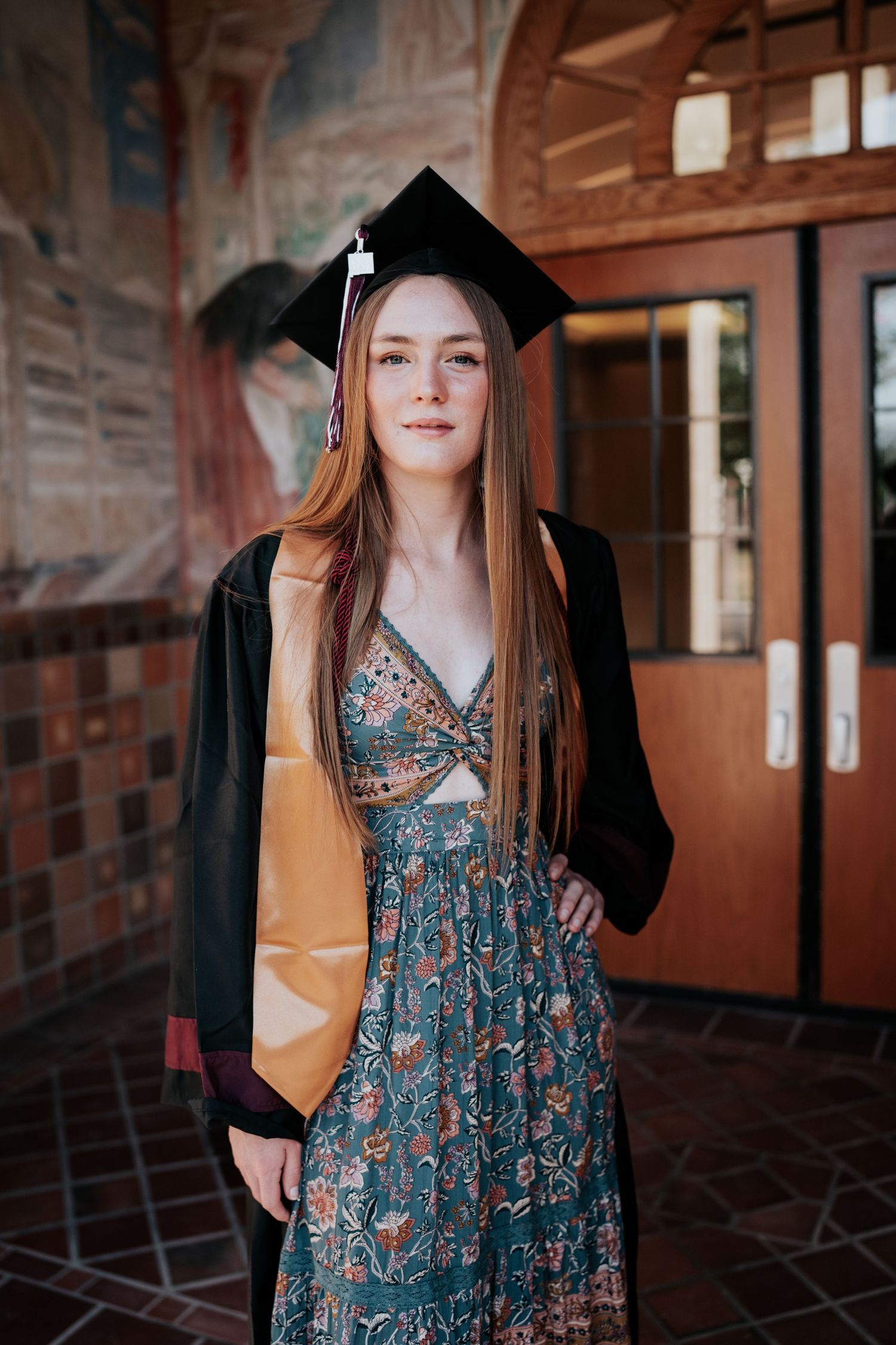 Female graduate in black cap and gold stole wearing floral dress posing at college campus.