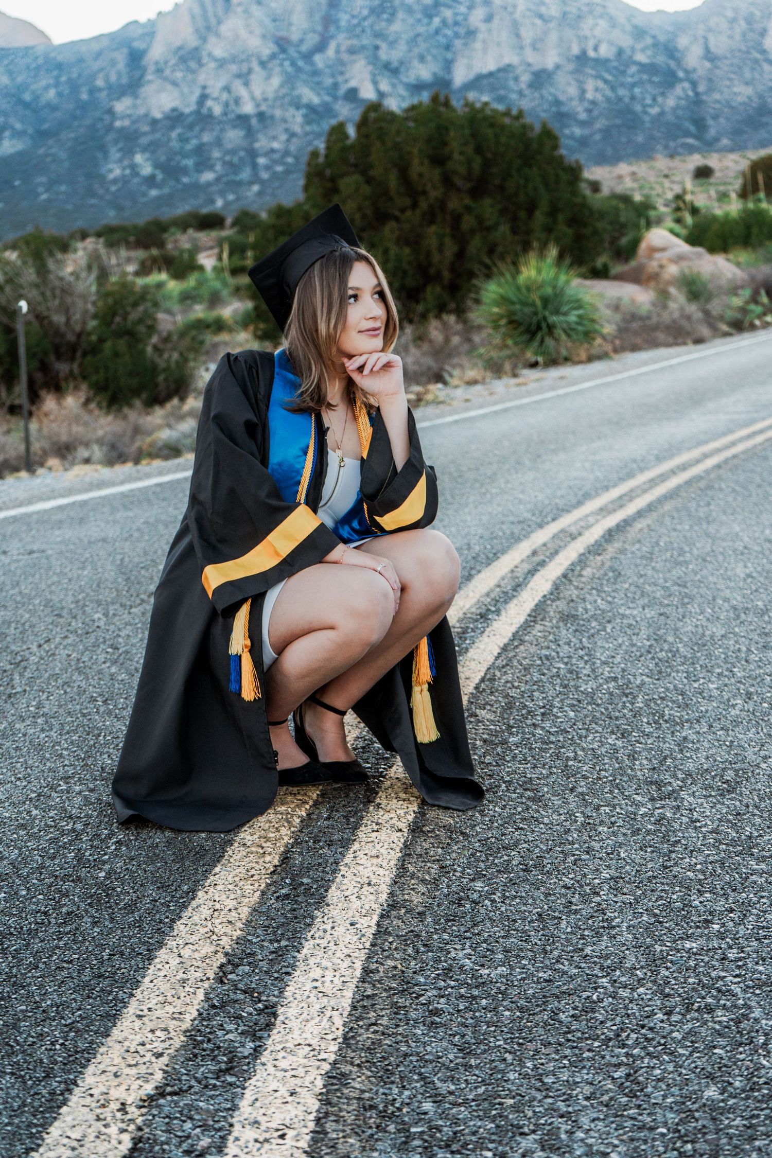 Graduate in black cap and gown crouching on a mountain road for a scenic graduation photo.