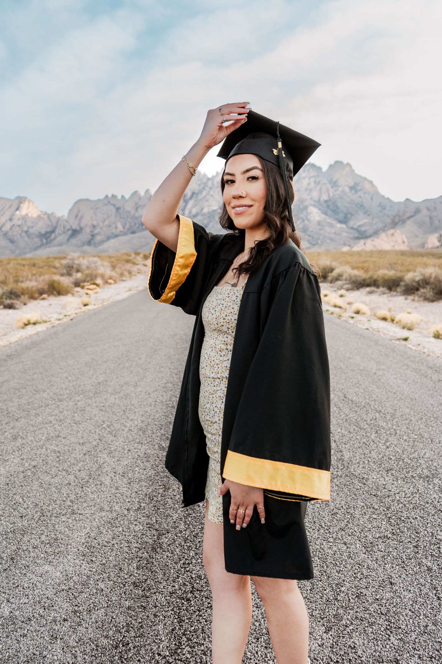 Female graduate in black and gold cap and gown standing on open road with mountain scenery.