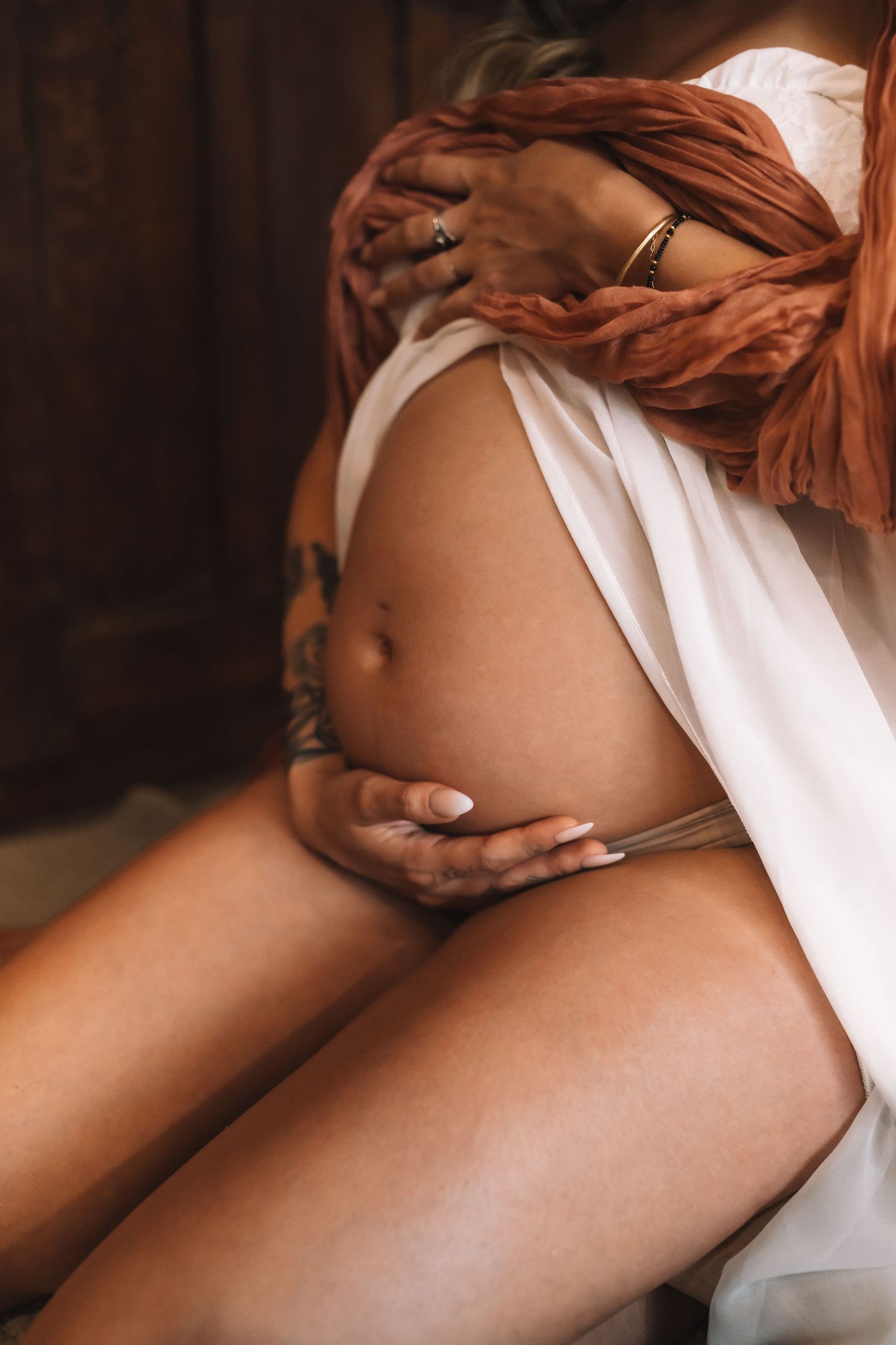 Close-up of pregnant belly with tattooed arm, gold bracelet, and terracotta scarf in artistic maternity photography.
