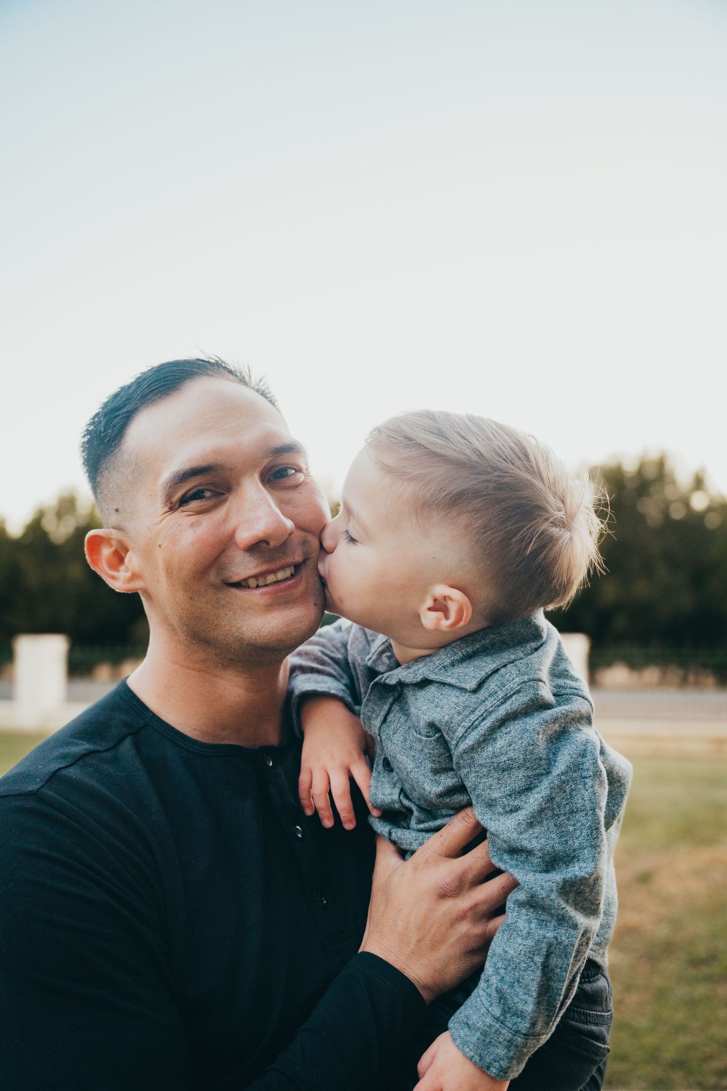 A smiling father holds his young toddler son who gives him a sweet kiss on the cheek outdoors.
