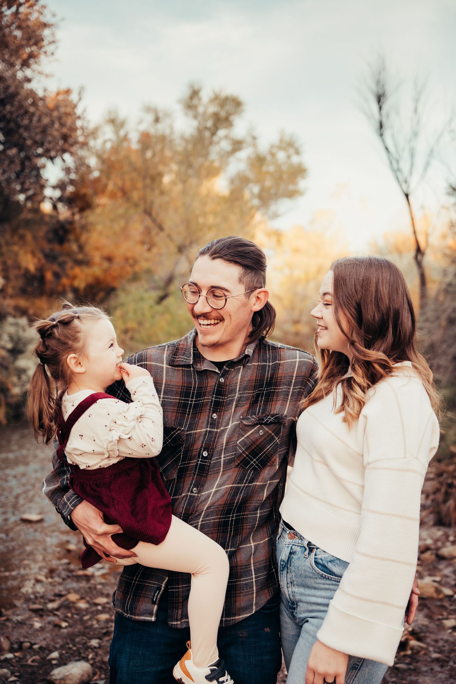 Happy family of three laughing outdoors in autumn, father holding toddler girl in burgundy dress with smiling mother beside him.