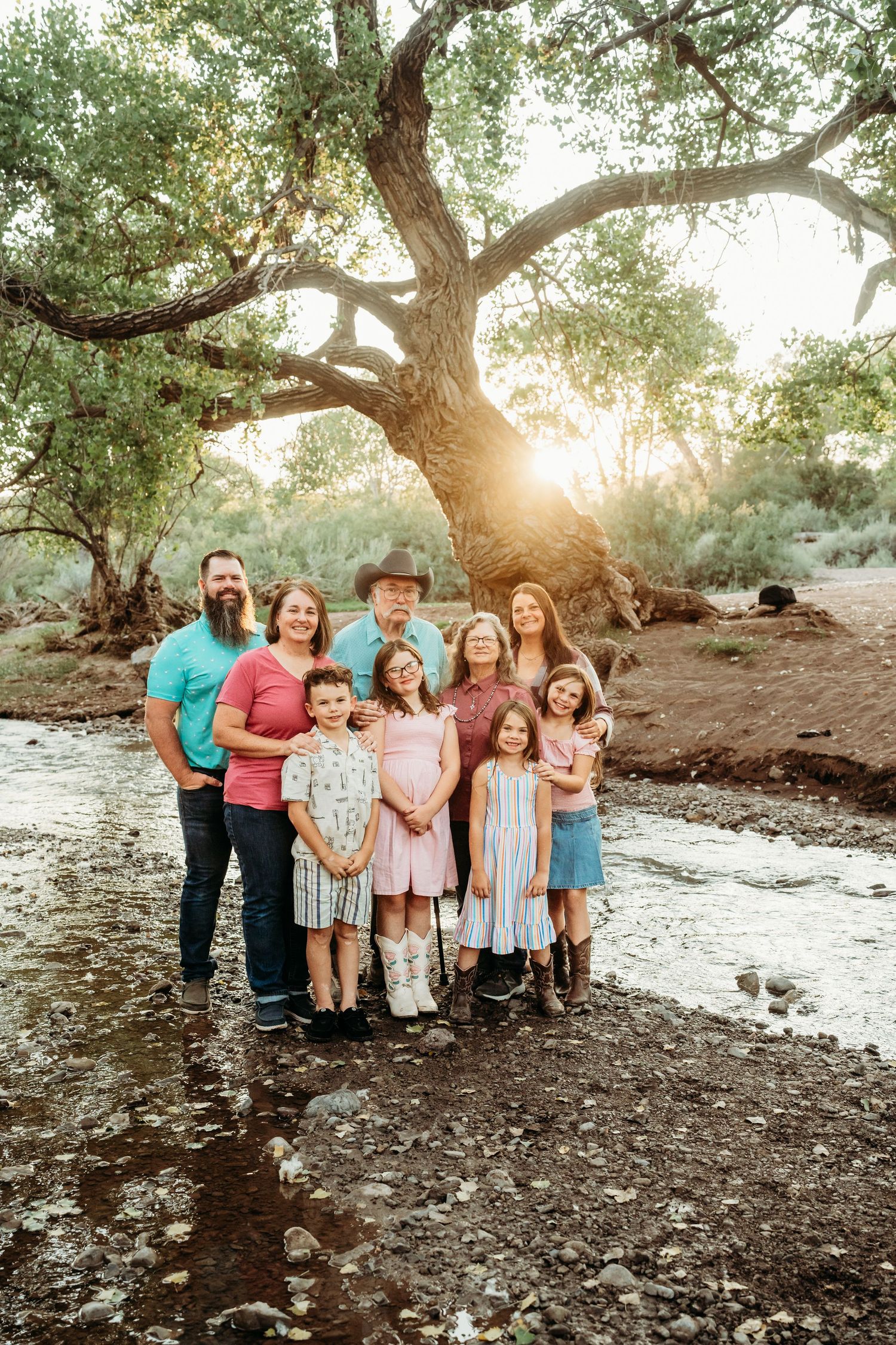 Extended multigenerational family posing by a sunlit creek under a large tree at golden hour.