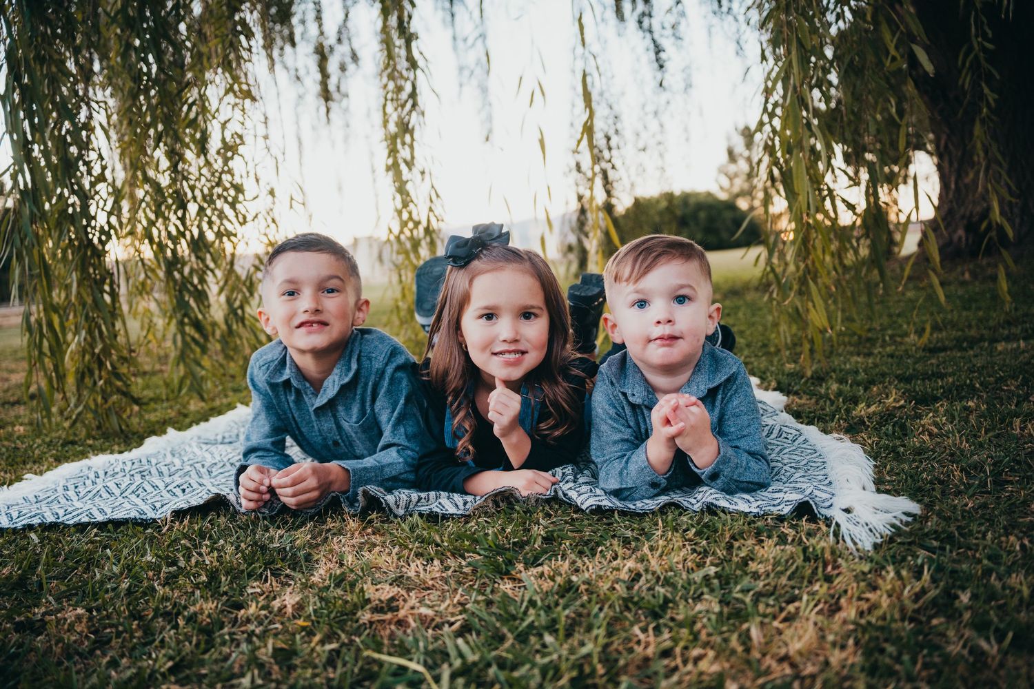 Three young siblings lie on a blanket in the grass beneath a weeping willow tree, smiling at the camera.