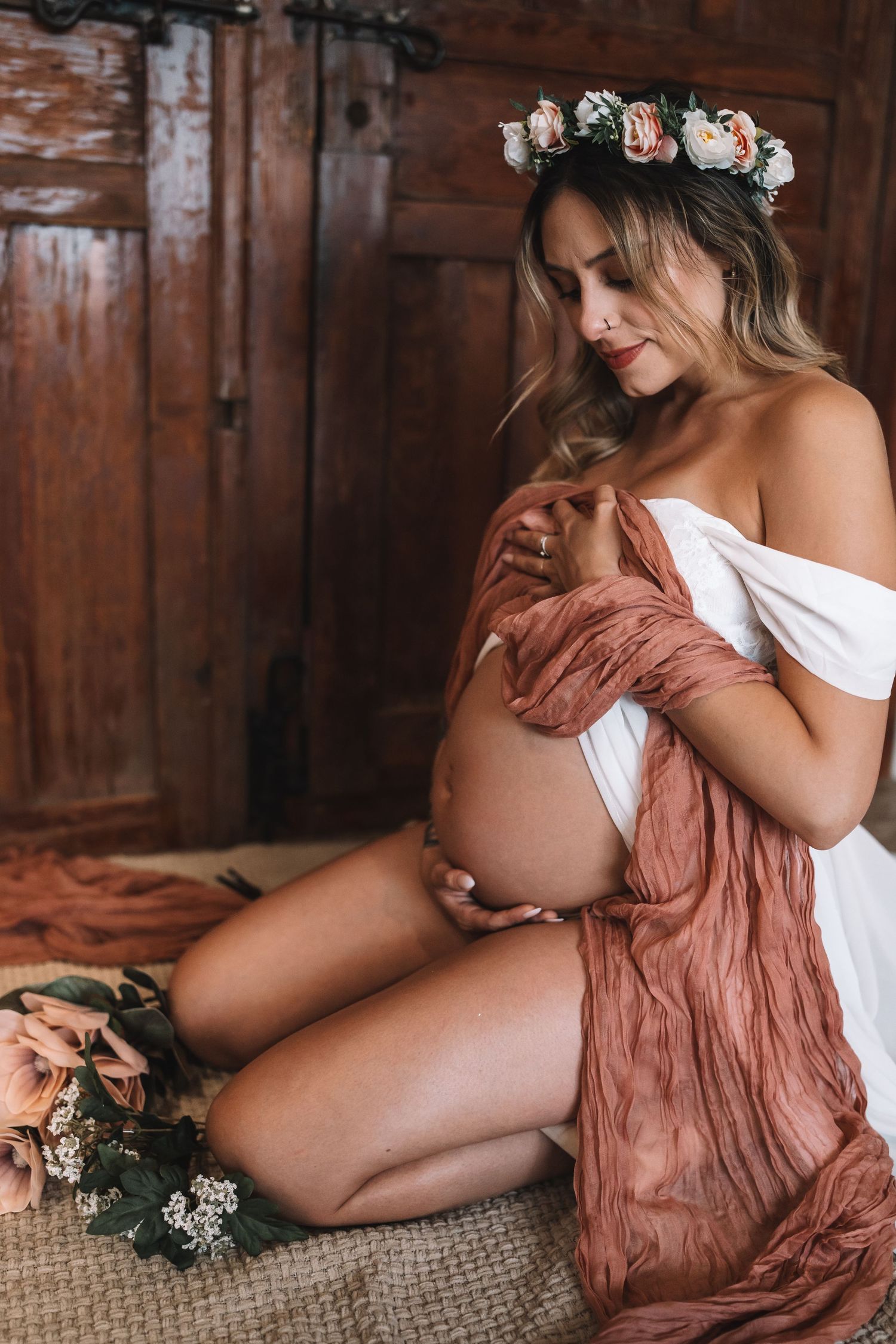 Beautiful pregnant woman wearing flower crown and white dress tenderly cradles her baby bump in boho maternity shoot.