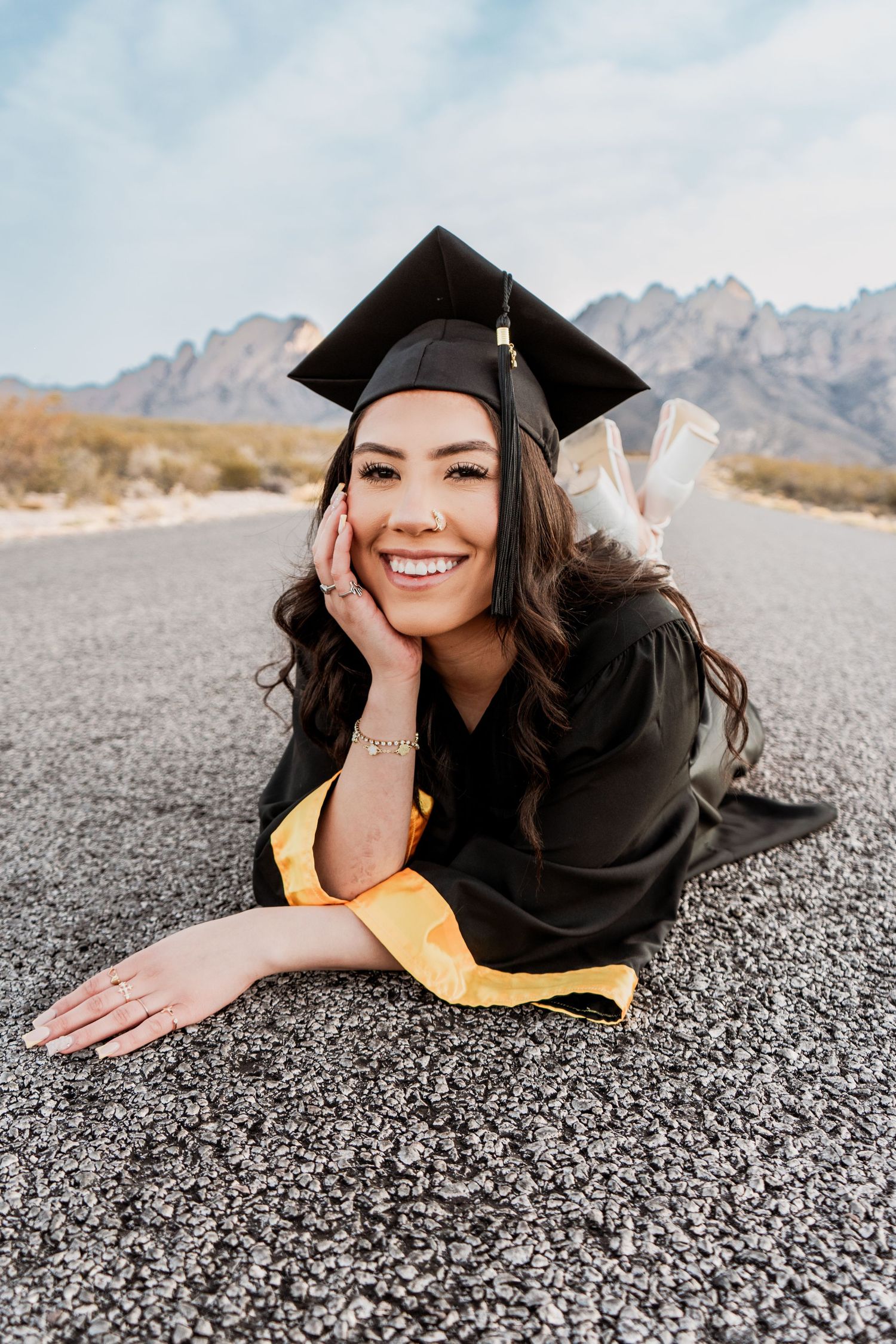 Smiling female graduate in black cap and gown lying on desert road with Organ Mountains backdrop.