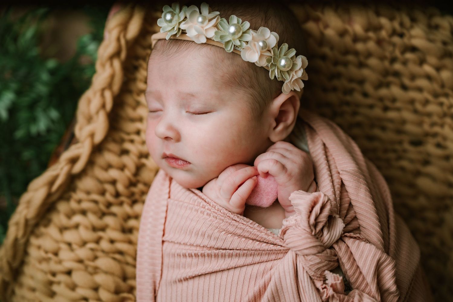 Peaceful newborn girl swaddled in pink wrap wearing white flower crown sleeping in basket.