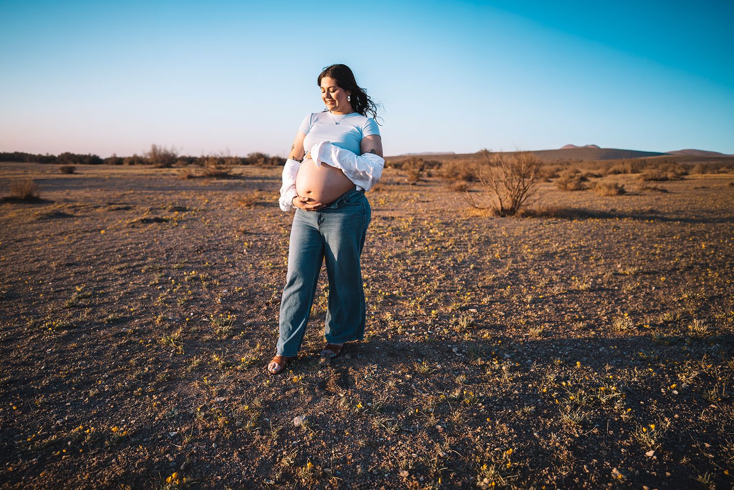 Pregnant woman looks down at baby bump standing in vast desert at sunset.