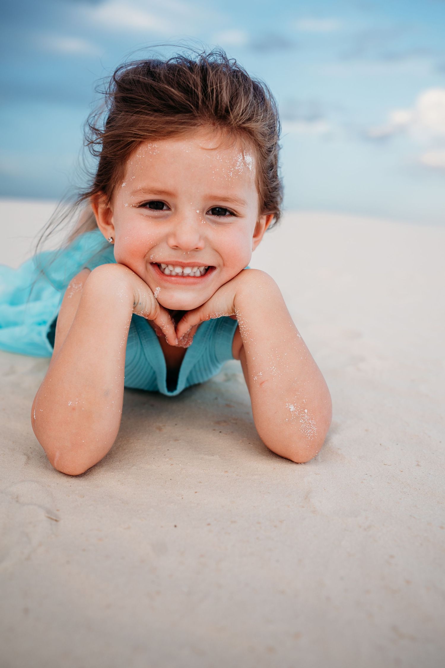 Adorable little girl in turquoise dress lying on white sand, smiling with hands under her chin.