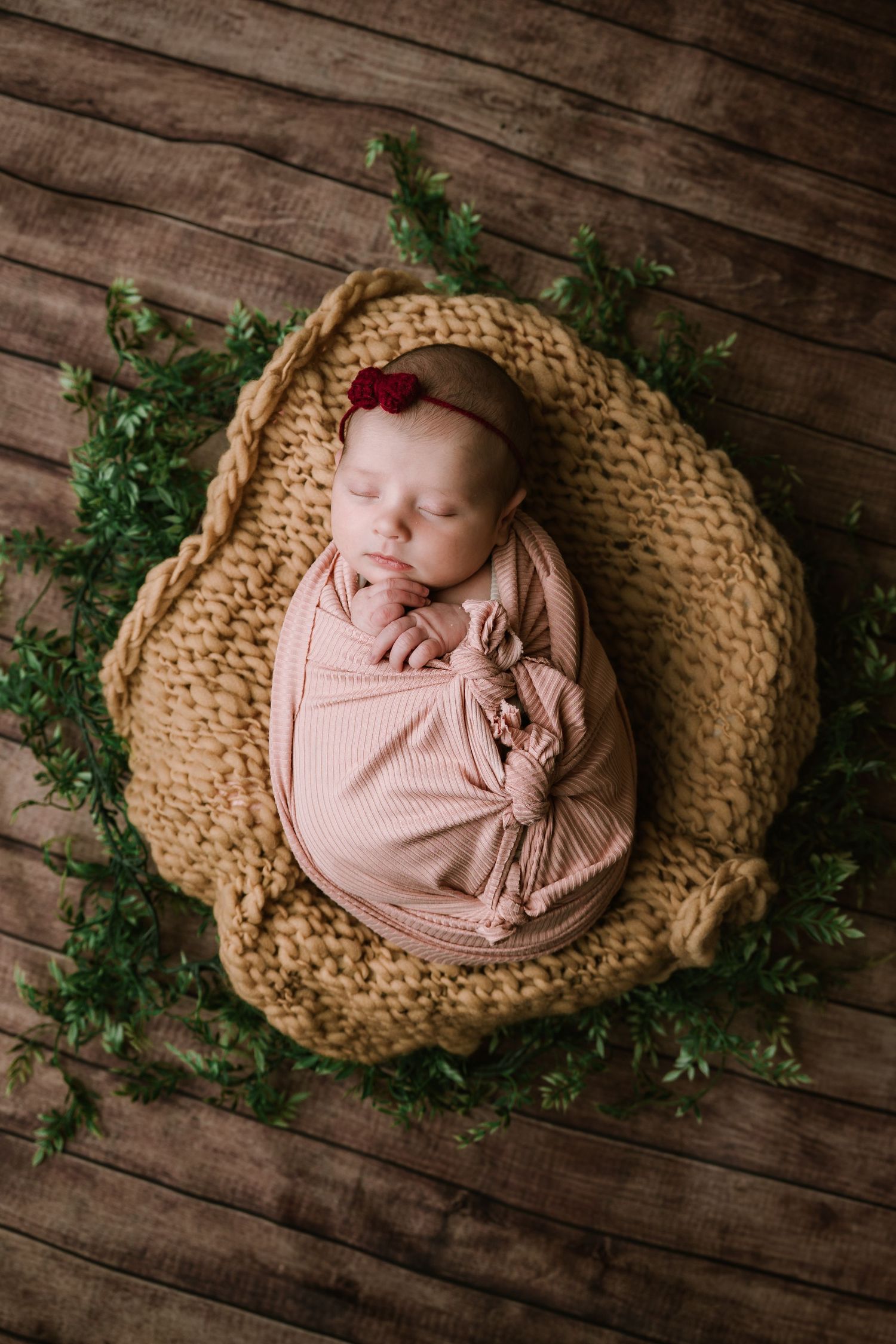 Newborn baby swaddled in pink wrap with red flower headband, sleeping in knit basket surrounded by greenery on wood floor.
