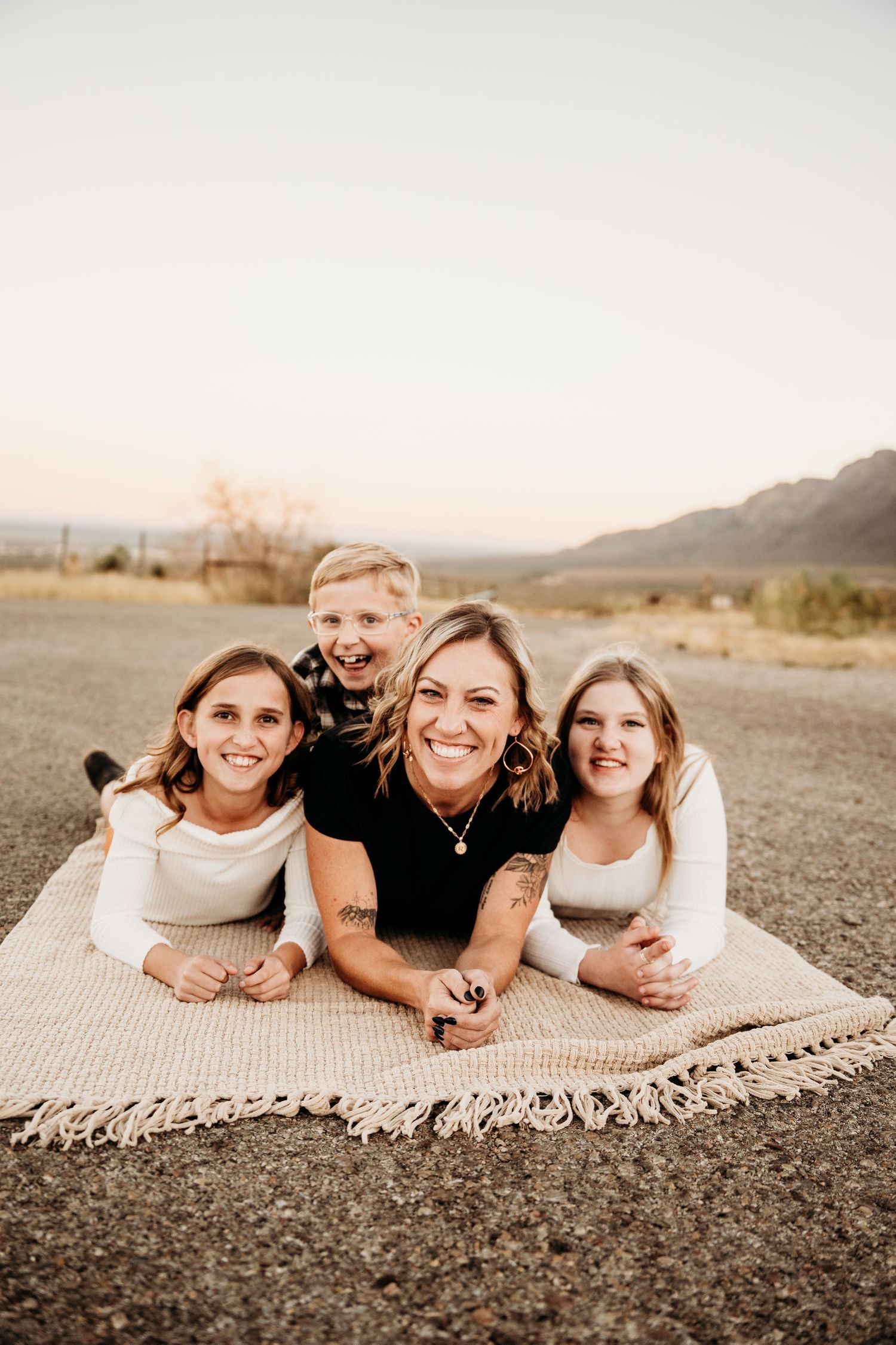 Smiling mother and three children lying on a blanket outdoors at golden hour with mountain backdrop.