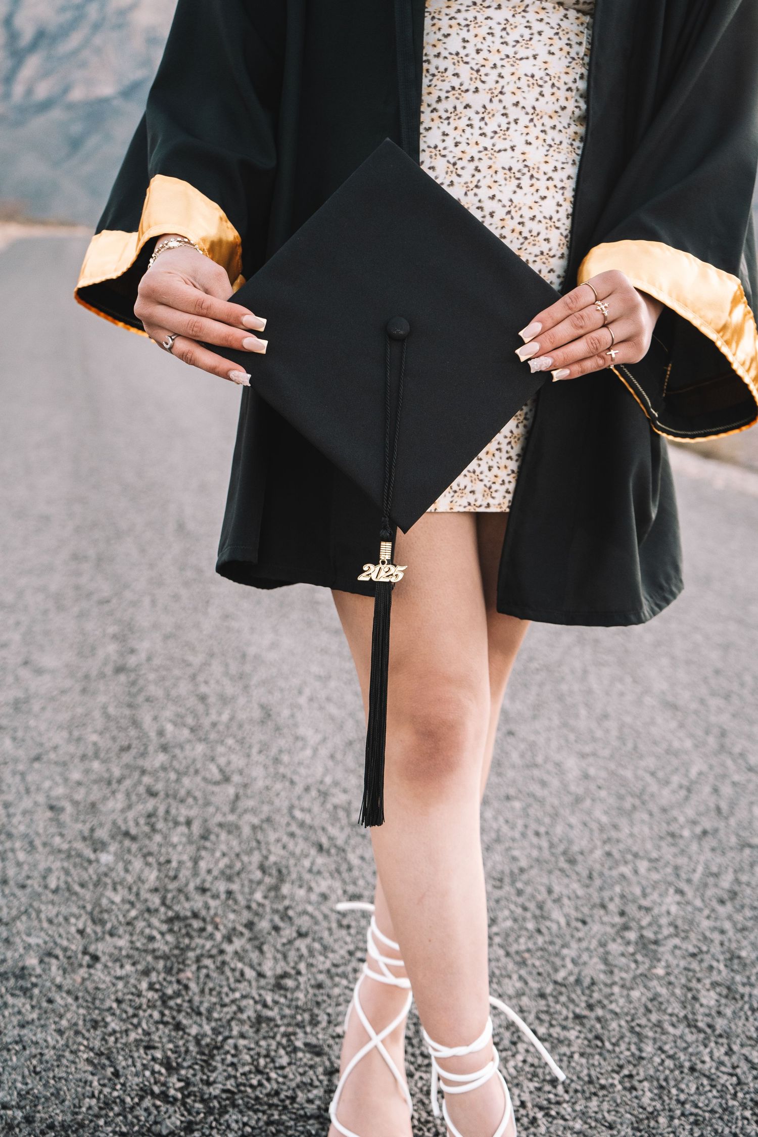 Close-up of a graduate in black and gold gown holding a 2025 mortarboard cap while wearing white heeled sandals.