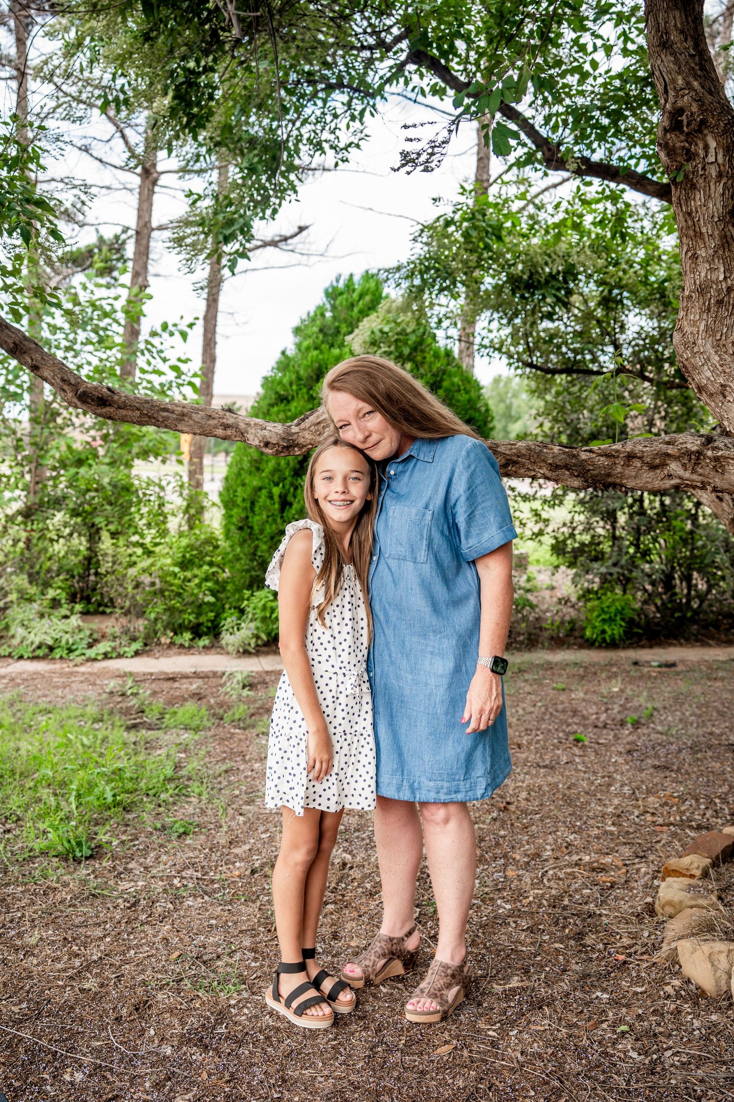 Two young girls in casual dresses pose together smiling under a large tree in an outdoor setting.