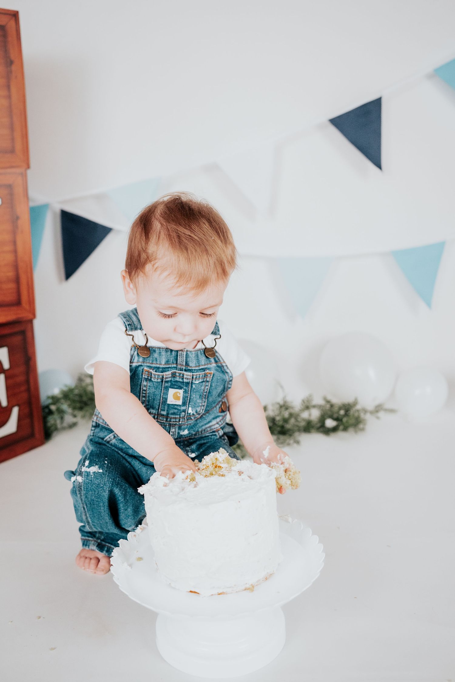Baby boy in Carhartt denim overalls digging into white frosted first birthday cake with blue banner backdrop.