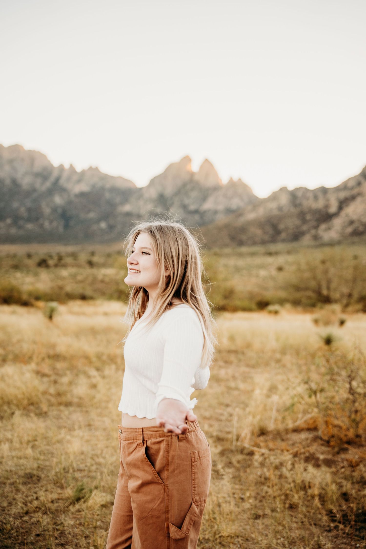 Teenage girl in white top and rust pants stands in sunlit desert field with mountain backdrop.