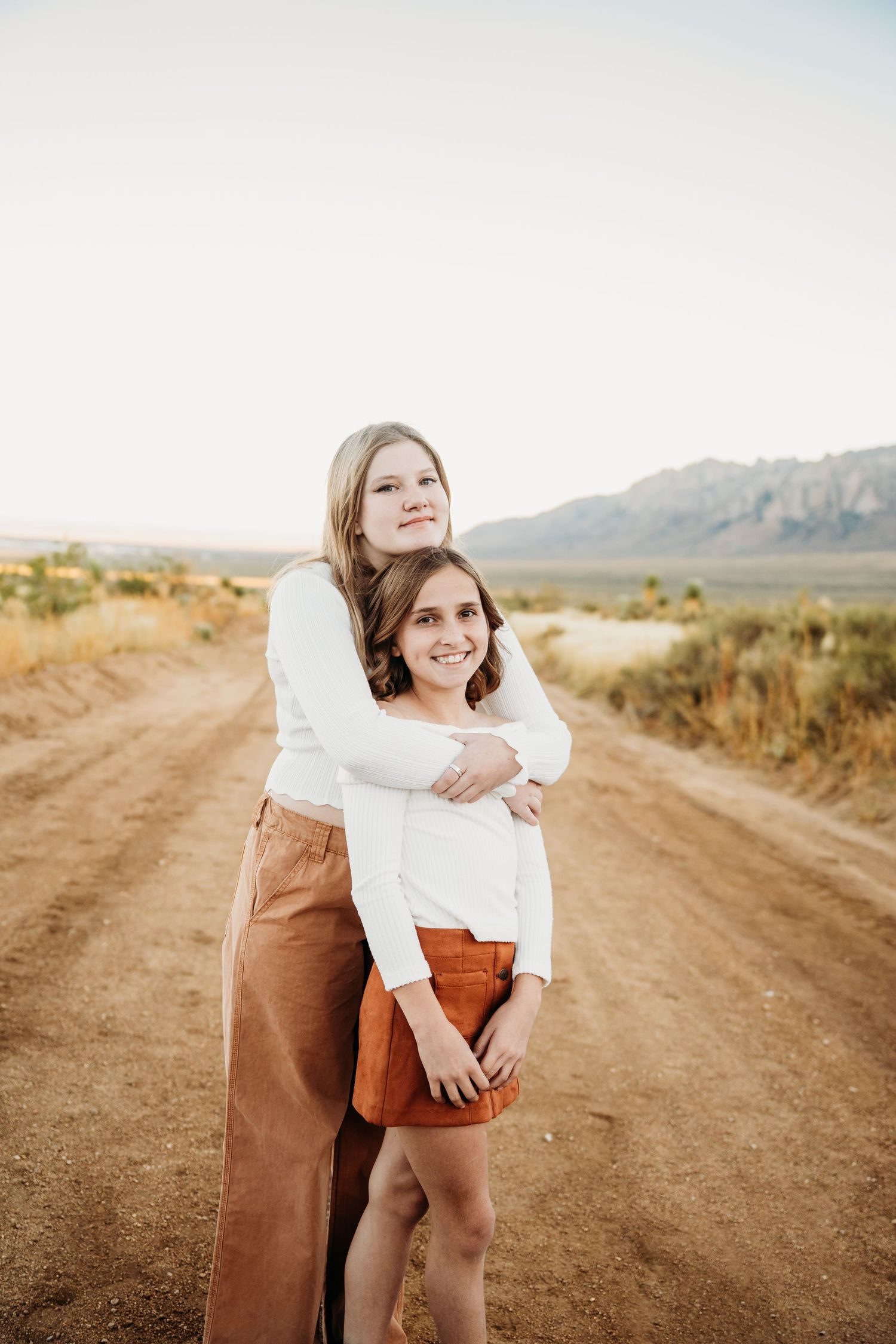 Two girls embrace warmly on a desert dirt road with mountains behind them.