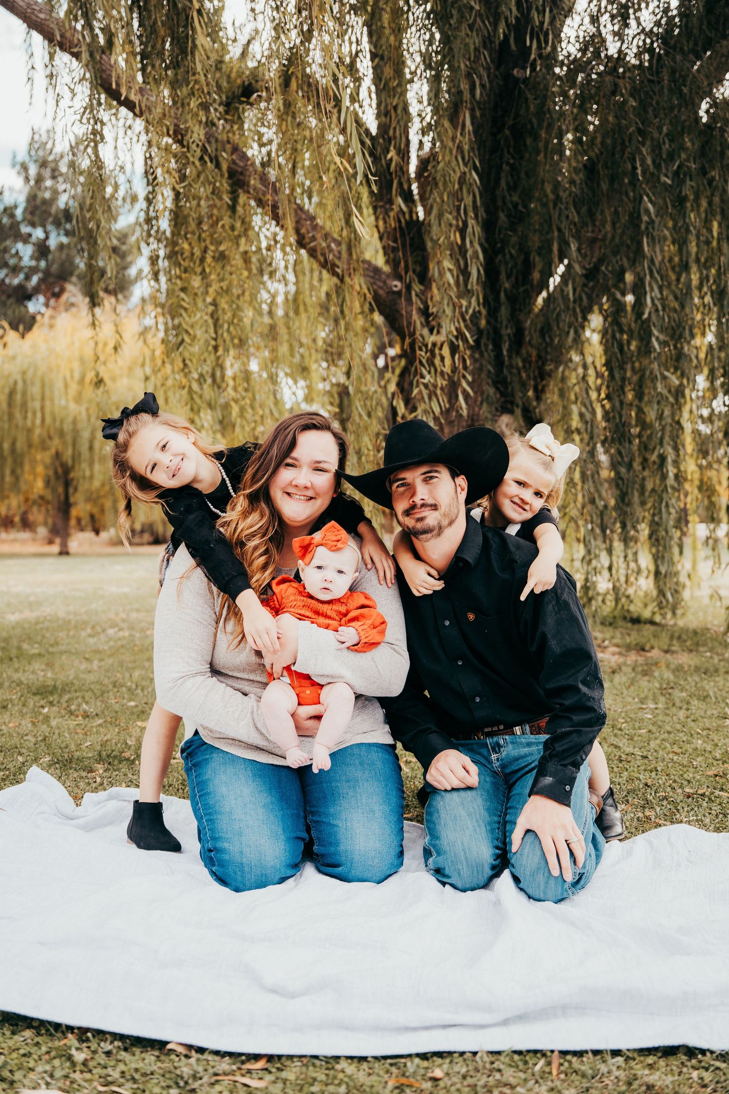 Smiling family with three children sitting on blanket under weeping willow tree in autumn park.