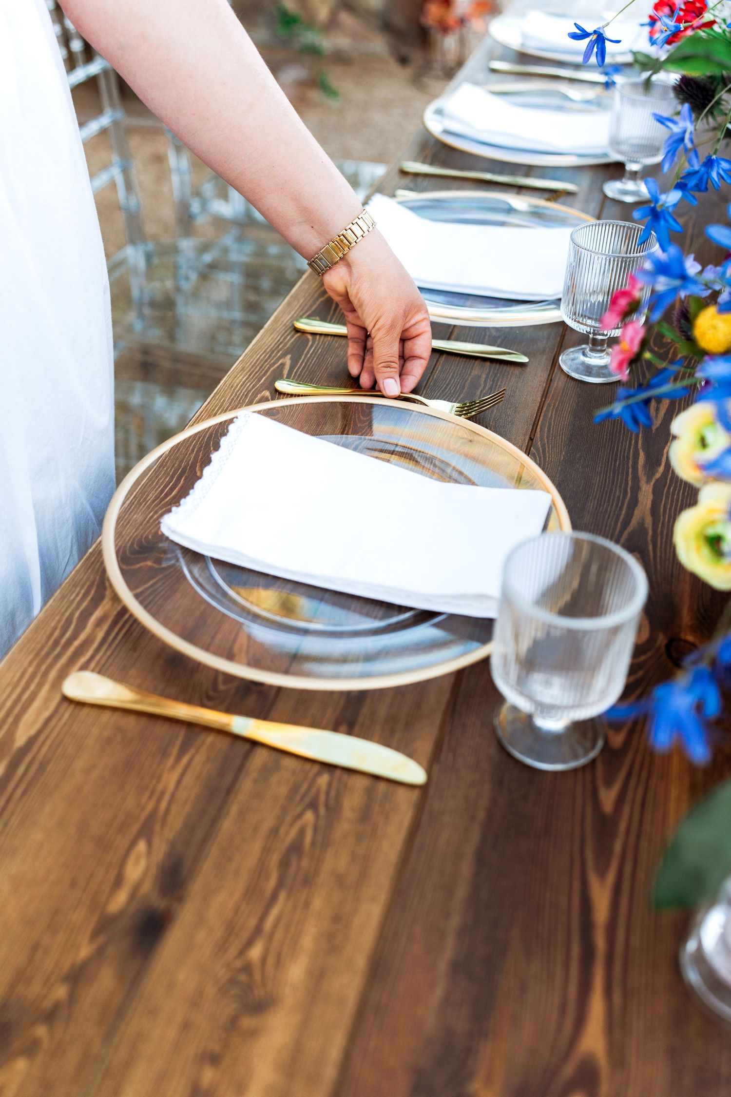Close-up of hand placing white linen napkin on gold-rimmed charger plate at an elegantly set outdoor wedding table.