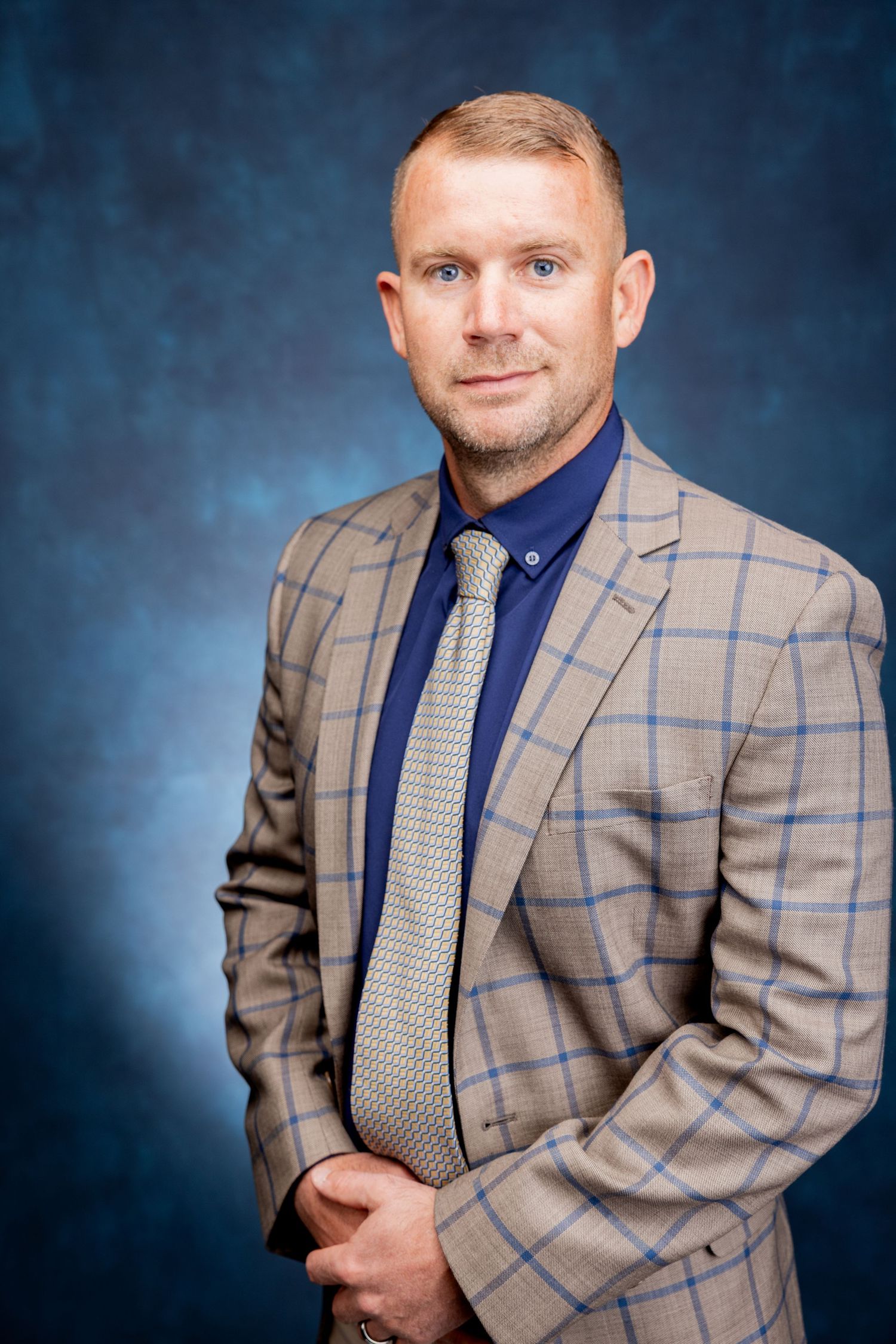 Professional standing portrait of a man in a tan plaid blazer, blue shirt, and gray tie on blue background.